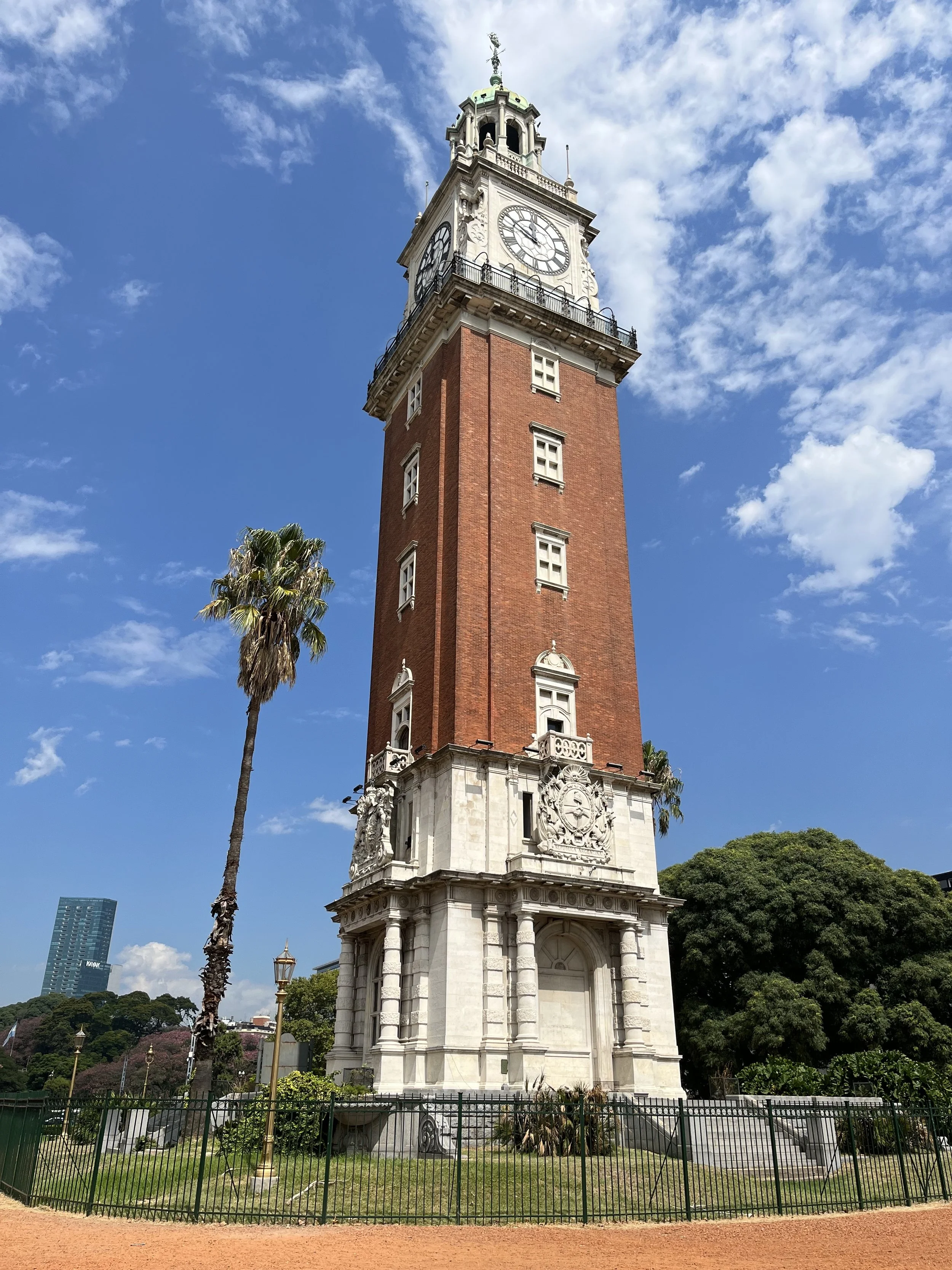 A tall clock tower with a white face and black Roman numerals, topped with a decorative cupola, against a partly cloudy blue sky. Palm trees and greenery surround the base of the tower.