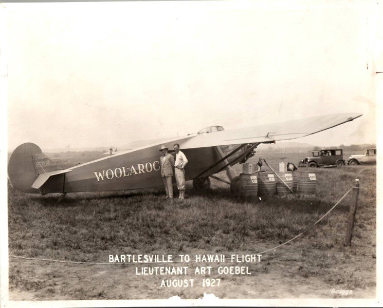 An old black-and-white photograph of a wooden airplane named Woolaroc on a grassy field with two men standing beside it, tanks, and vehicles in the background, dated August 1927, from Bartlesville to Hawaii flight by Lieutenant Art Goebel.