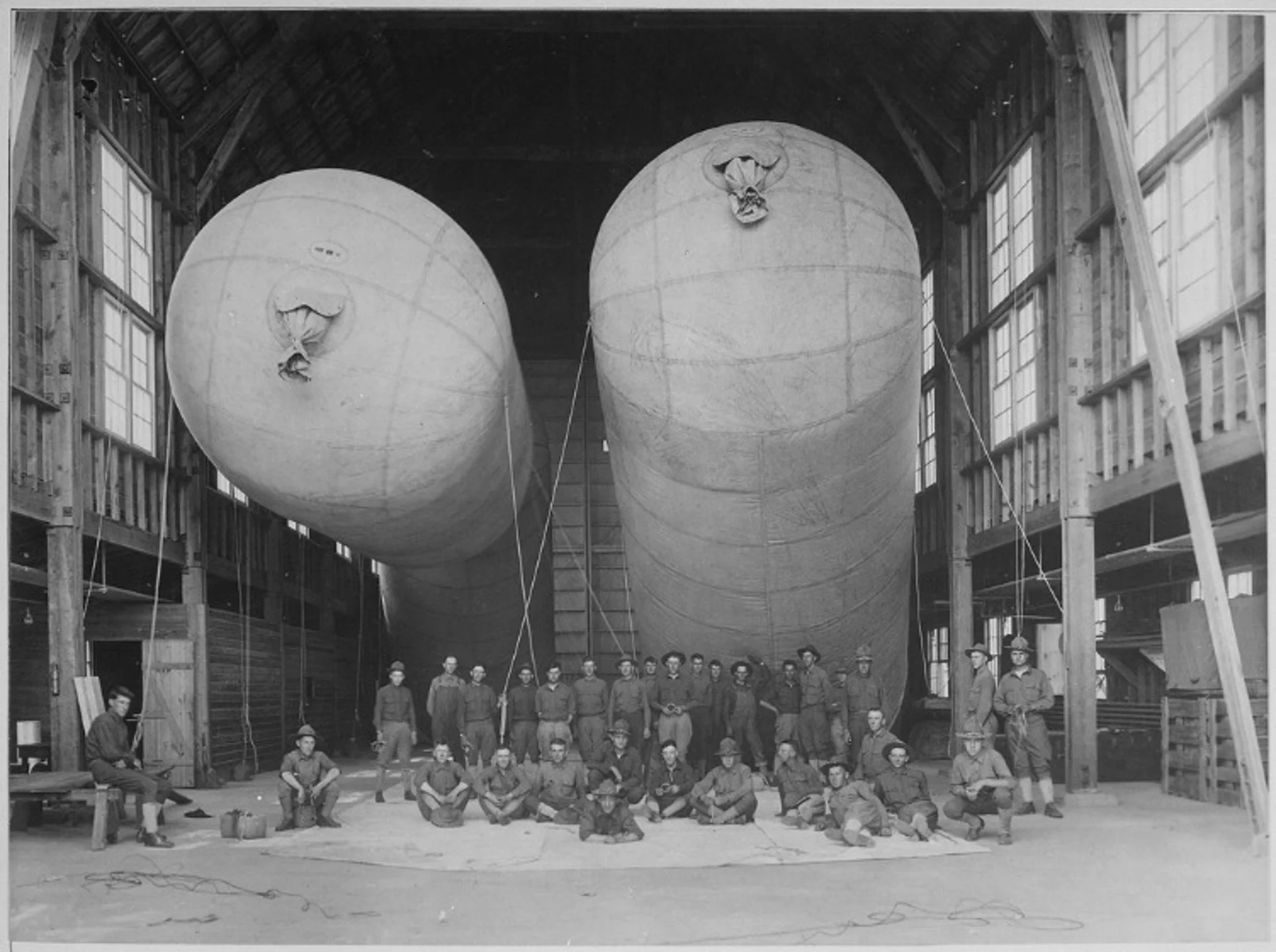 A group of men, some sitting and some standing, posing inside a large wooden hangar or workshop that contains two enormous spherical objects suspended in the air. The objects appear to be part of a large experimental or engineering project.