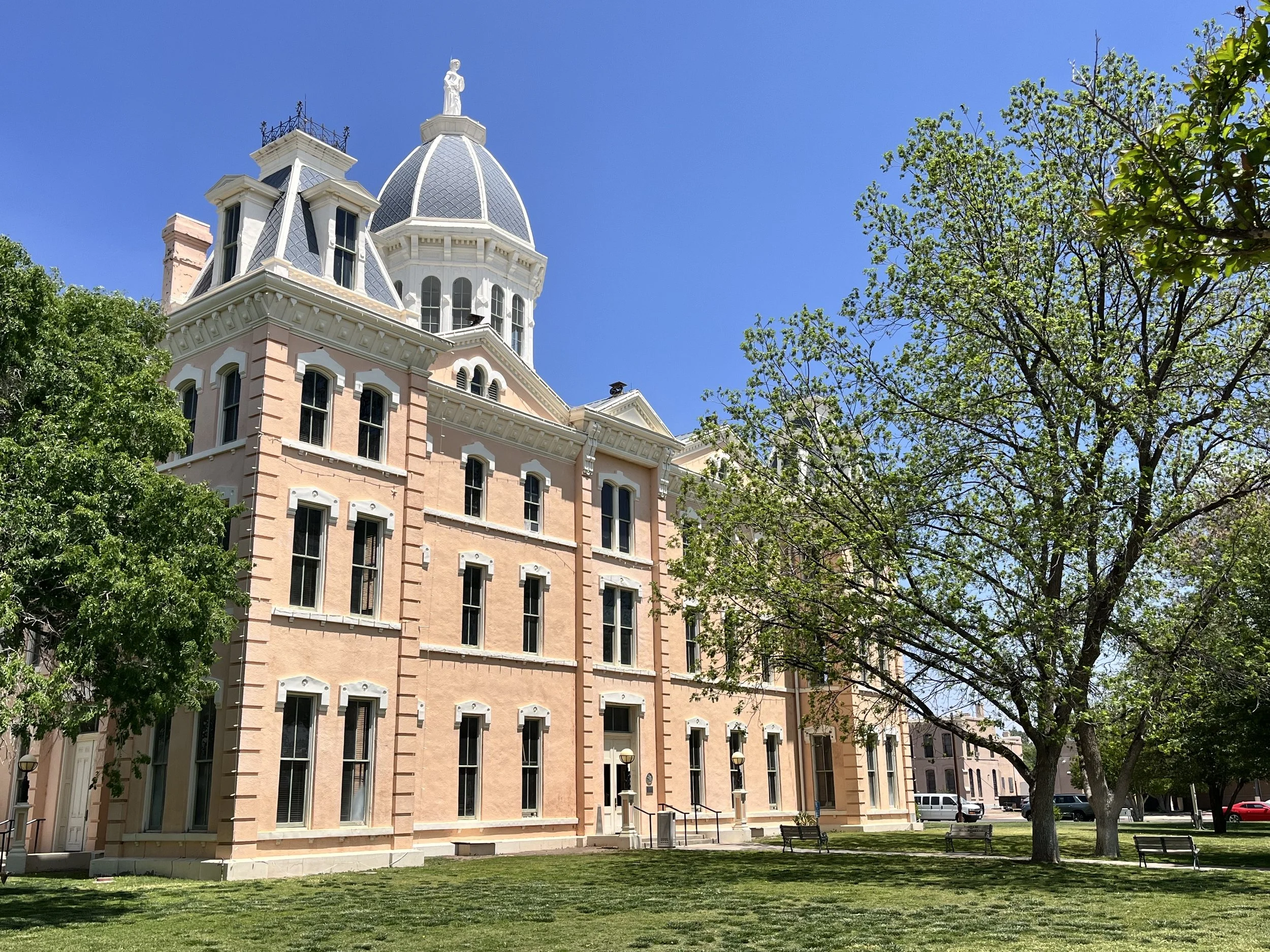 A large historic pink building with a domed tower and statue on top, surrounded by green trees and a park with benches, clear blue sky in the background.