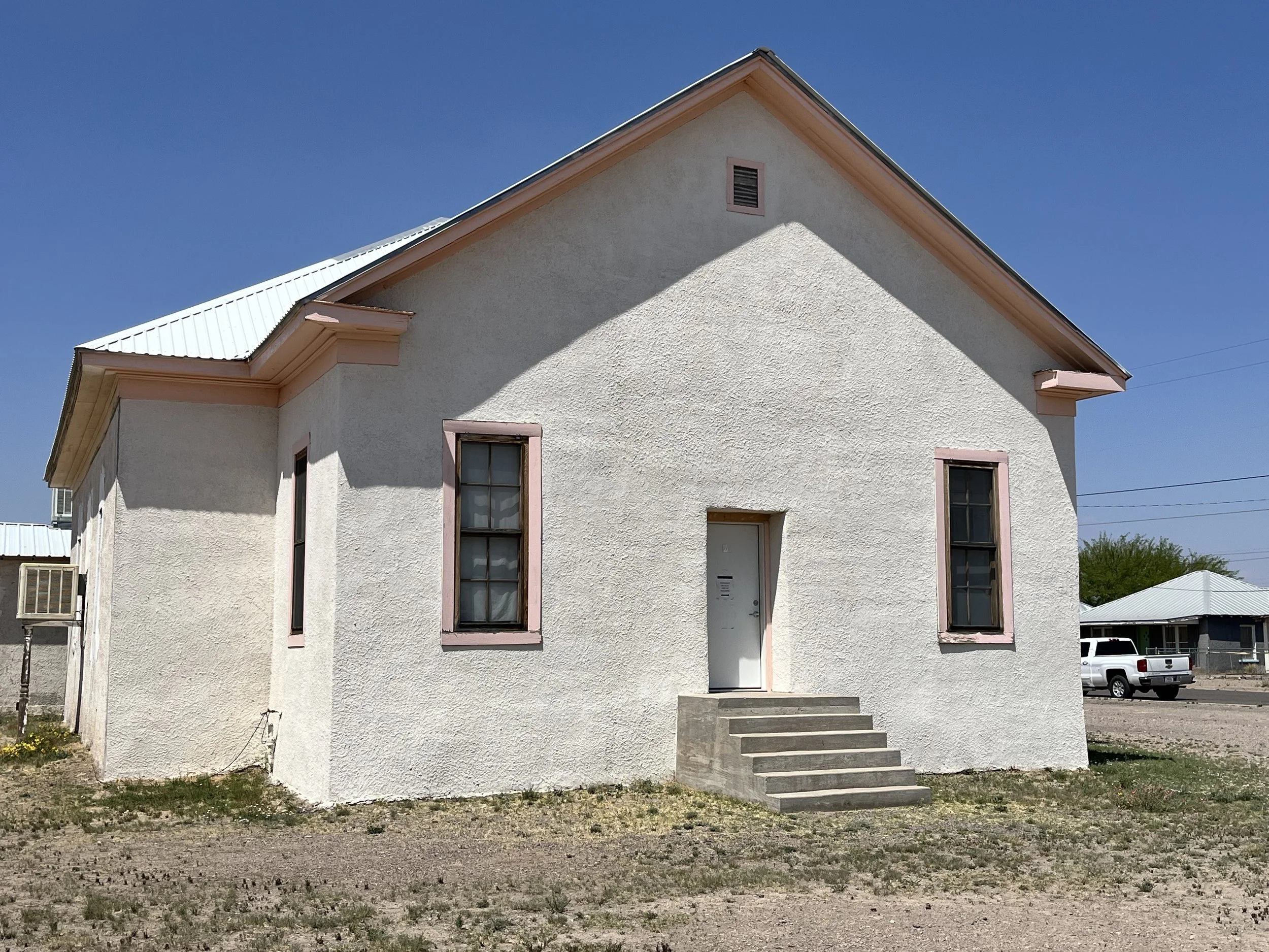 A white two-story house with pink window frames, a set of concrete steps leading to the front door, and a metal roof. The house is on a gravel lot with some grass, under a clear blue sky.