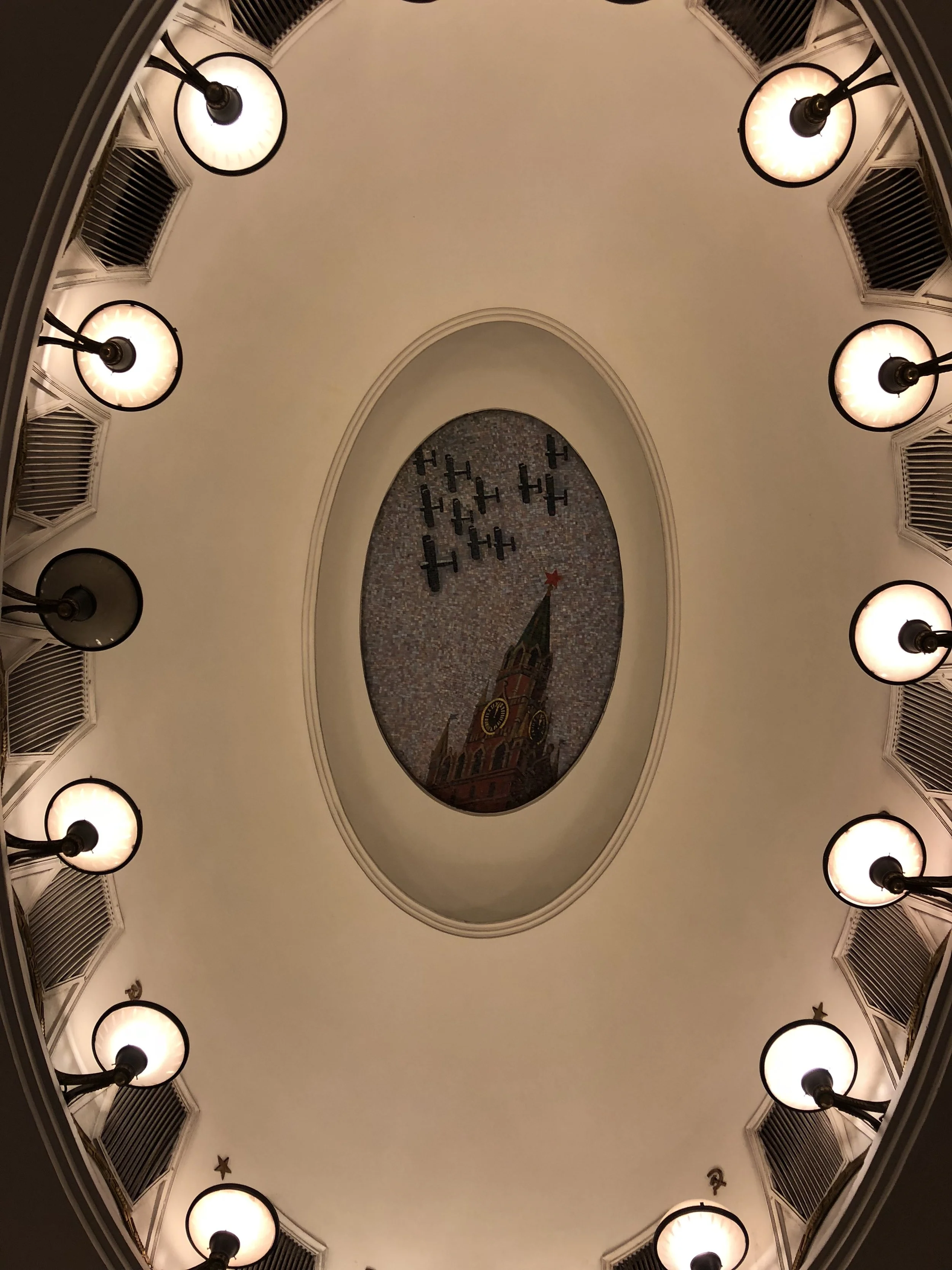 View of the ceiling with a mural of a clock tower and planes, surrounded by eleven evenly spaced lights.