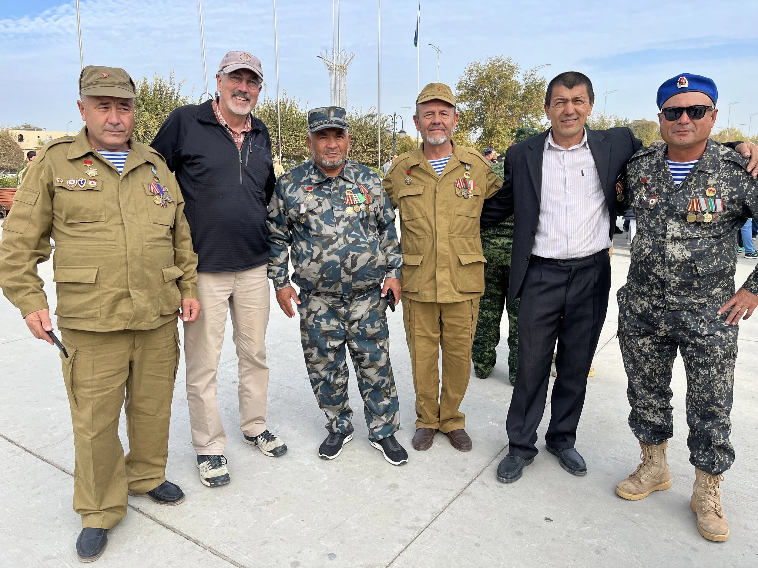 A group of seven men, some in military uniforms with medals, standing outdoors on a paved area, with trees and a clear sky in the background.