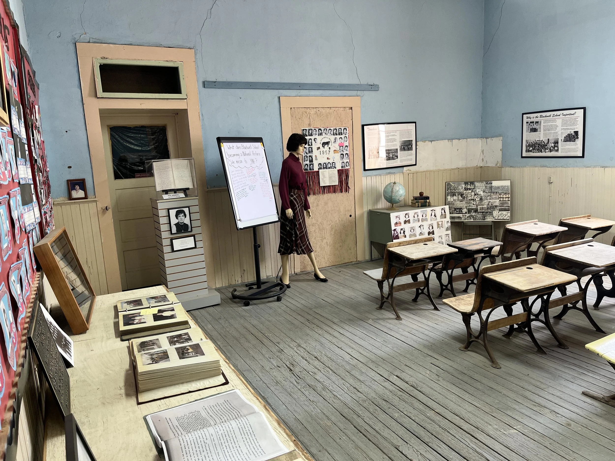 A vintage classroom with wooden desks and chairs, educational posters, photographs, a globe, and a mannequin dressed as a teacher. A whiteboard and display boards are also present.