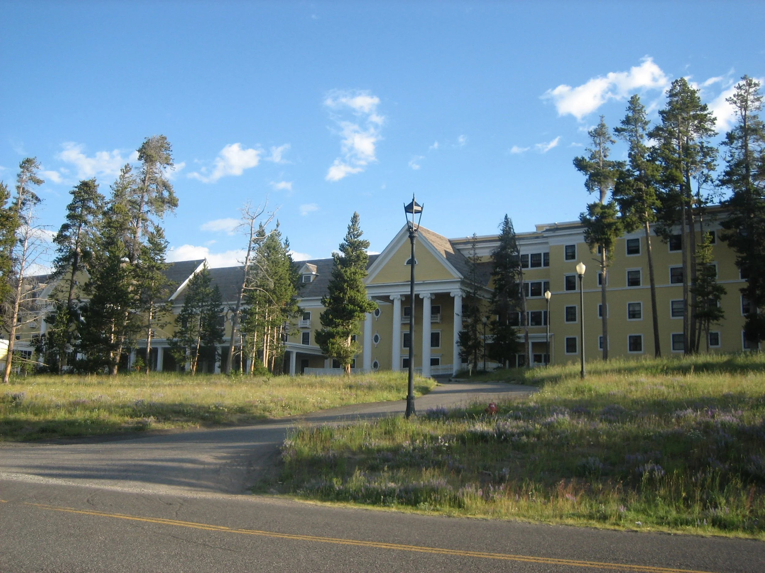 A large yellow hotel or apartment building with multiple floors, white columns at the entrance, surrounded by tall pine trees on a bright, partly cloudy day.