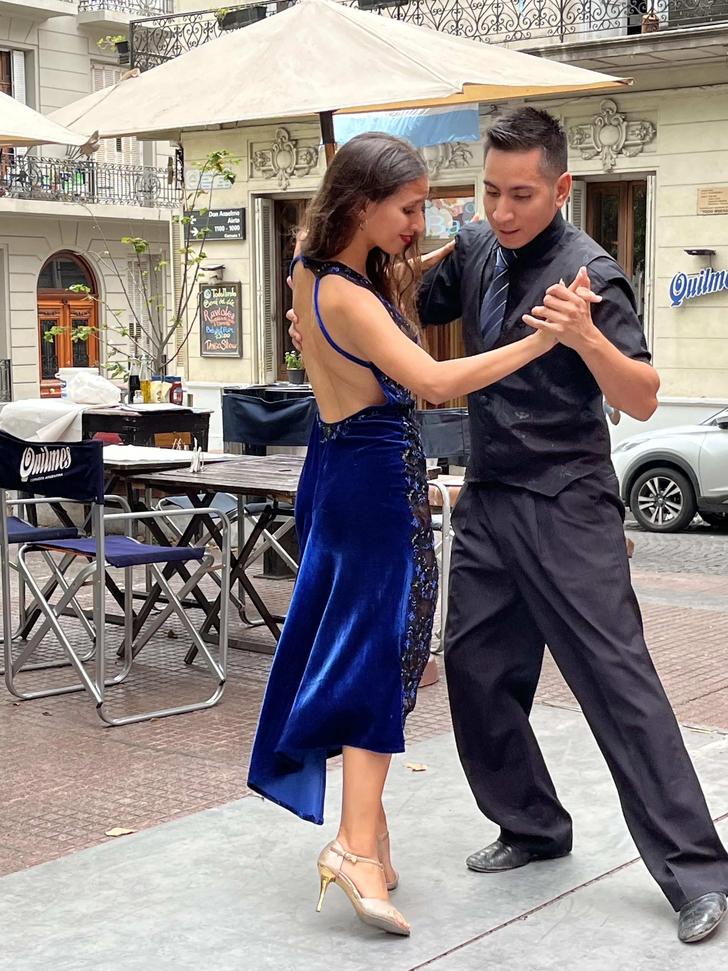 A man and woman are dancing outdoors on a city sidewalk. The woman is wearing a blue dress with an open back and tan high heels. The man is dressed in black with a striped tie. They are holding each other closely while dancing.