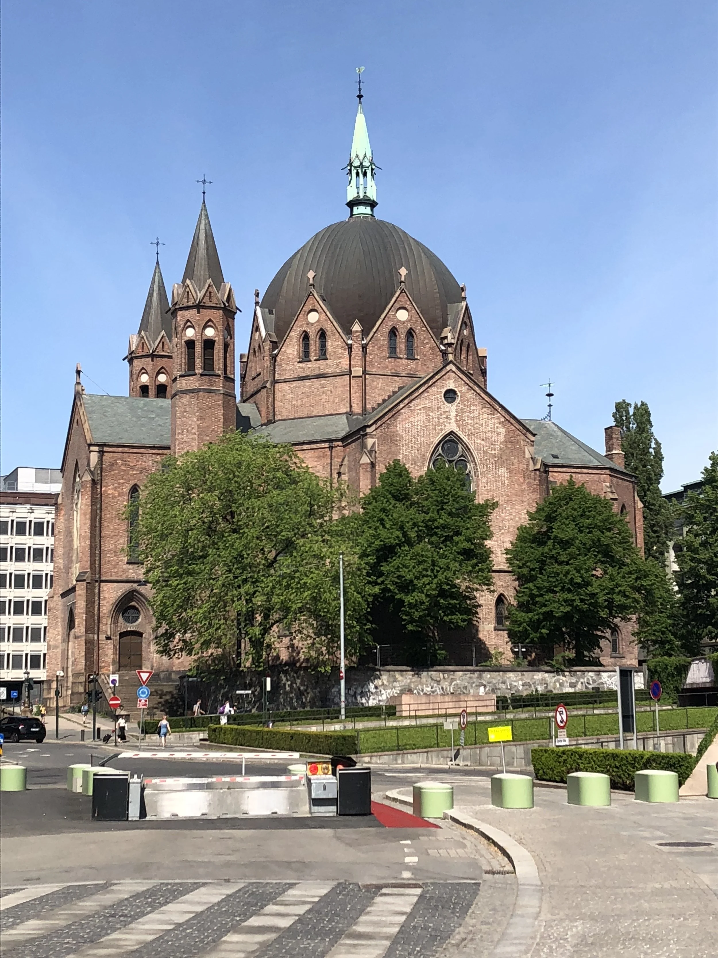 A large red brick church with a prominent black dome and tall spires, surrounded by greenery and situated in an urban area with traffic barriers and crossing markings.
