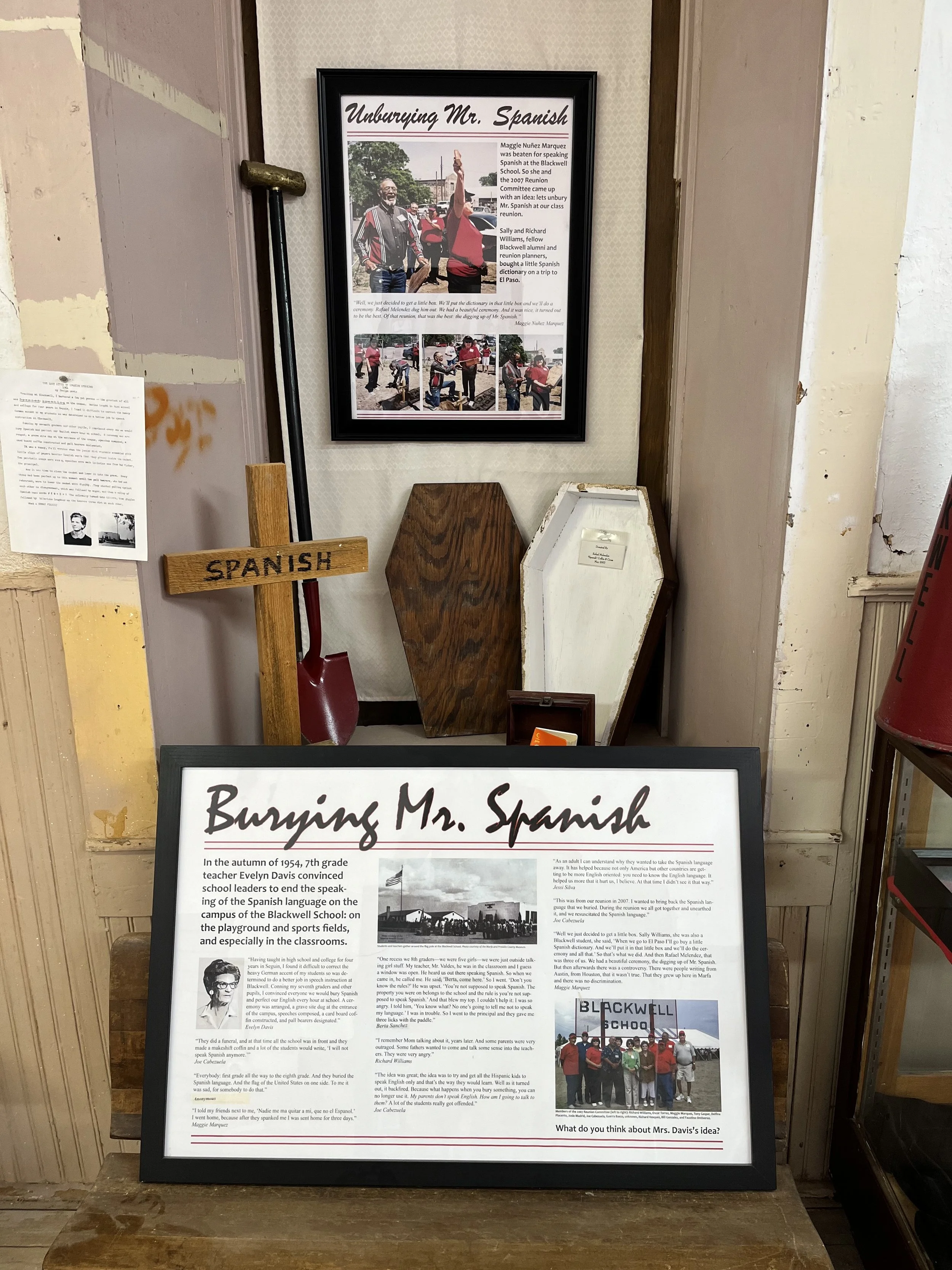 Display about the burial of Mr. Spanish with a framed article titled 'Burying Mr. Spanish,' along with several small photographs, wooden coffin-shaped objects labeled 'Spanish,' a shovel, and a small framed photo of a group of people in front of a sign that says 'Blackwell School.'