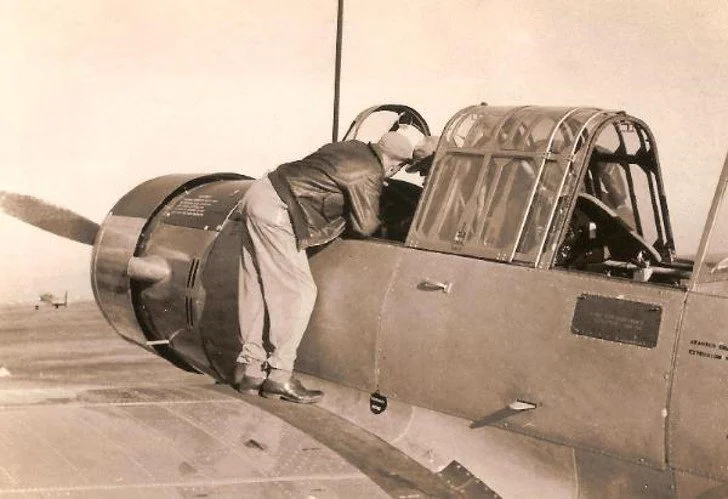 A man leaning into the cockpit of a vintage military jet aircraft on the ground, examining or working on the interior.