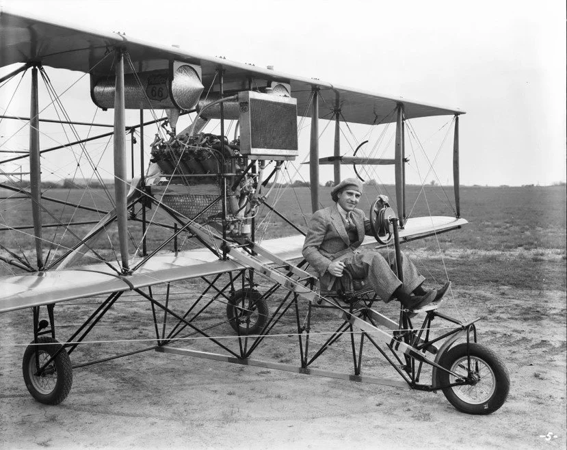 Black and white photo of a vintage airplane with a woman sitting in the cockpit, holding a control stick, on an open field.