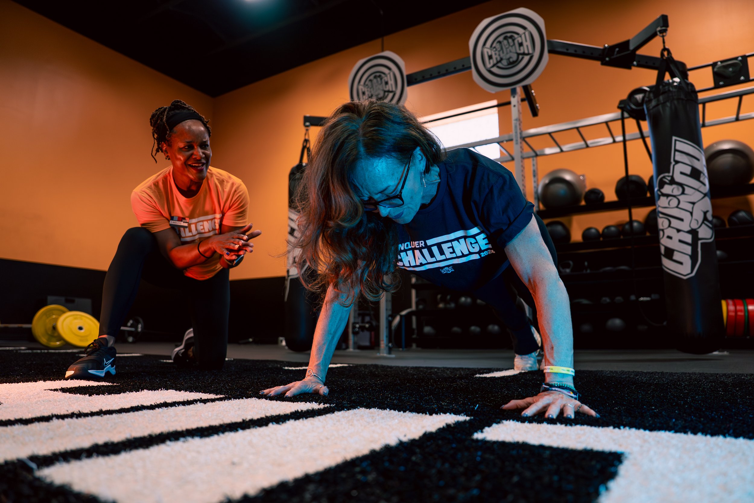 Two women in a gym, one is doing push-ups on a black and white striped mat, the other is mentoring or encouraging her. The gym has weightlifting equipment and bright orange walls.