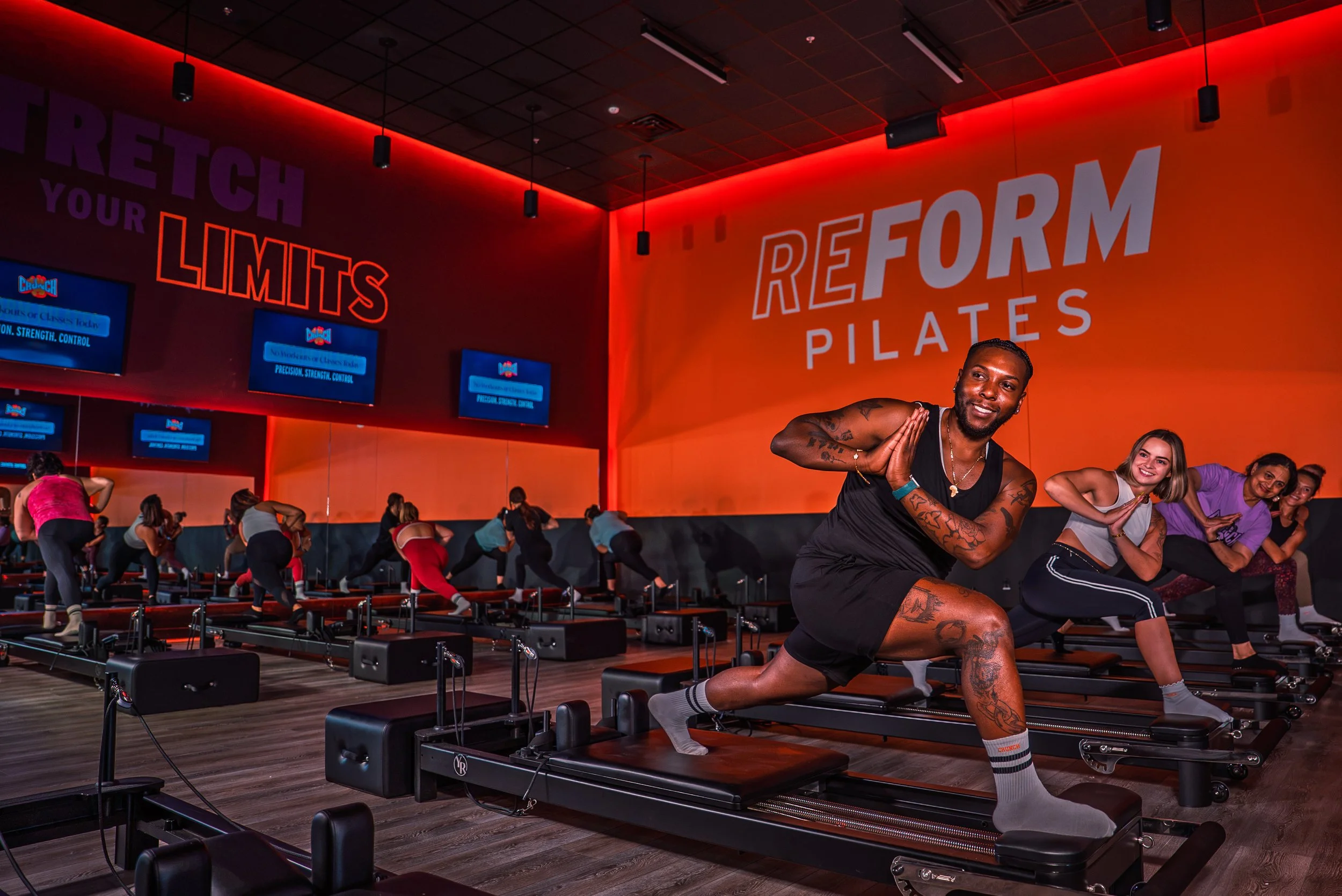A fitness class at a Pilates studio with participants on reformer machines performing exercises. The studio has dark flooring, orange walls with large white and purple text that reads 'REBUILD YOUR LIMITS' and 'REFORM PILATES'. A smiling man in a bla