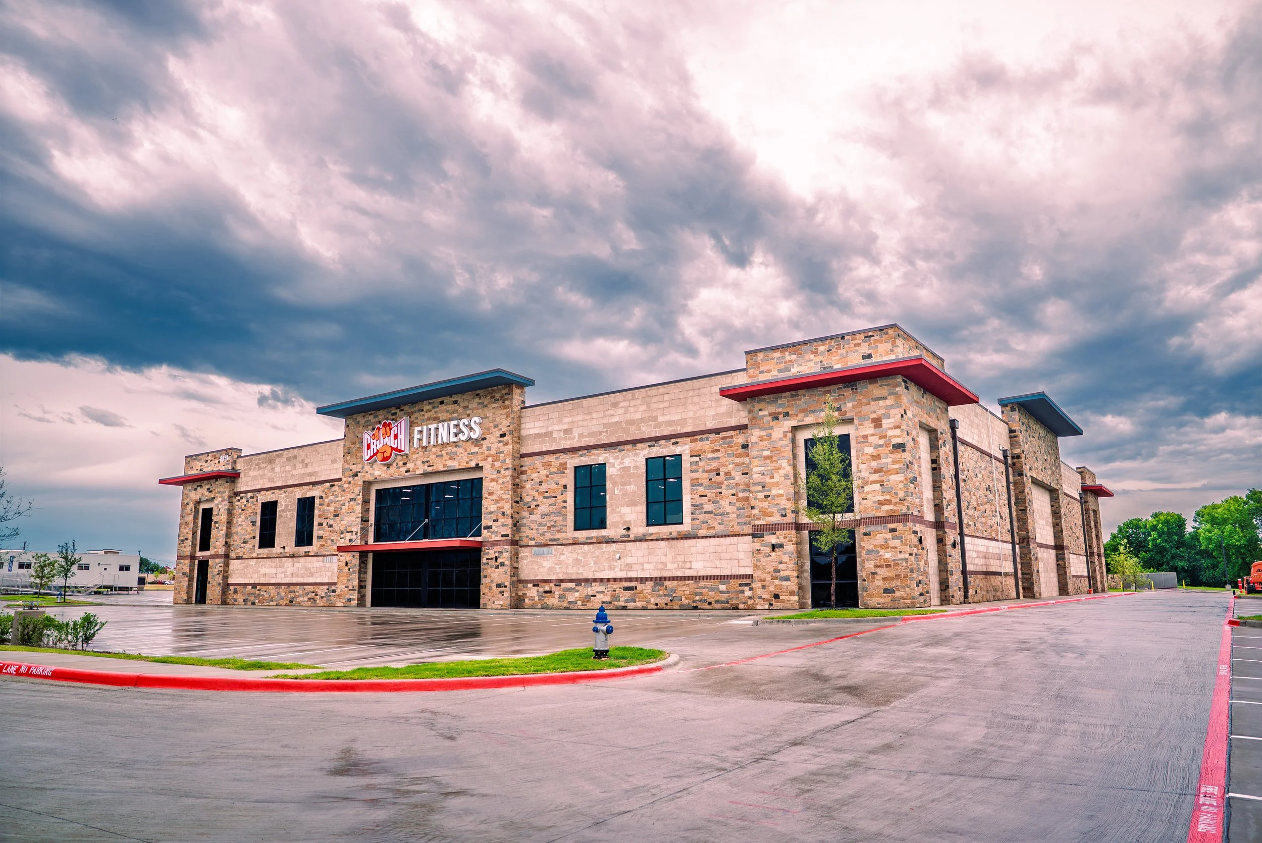 Empty parking lot in front of a modern brick and stone fitness center building with a cloudy sky overhead.