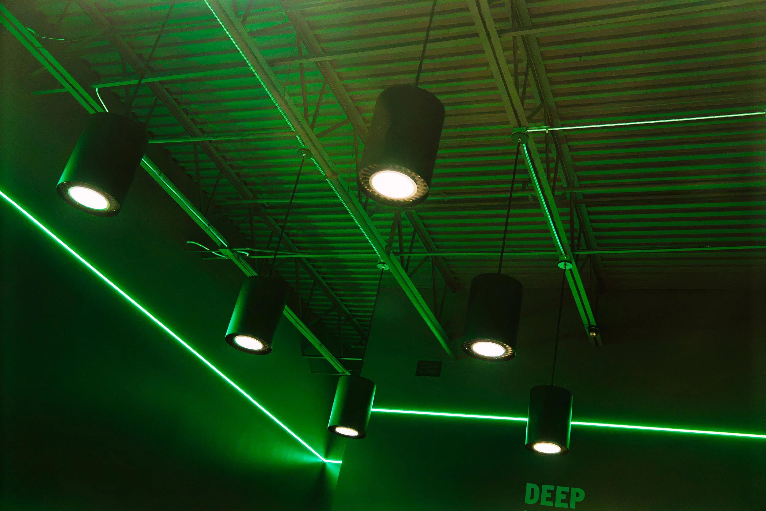 Black ceiling lights hanging against a ceiling with green neon lighting and metal beams.