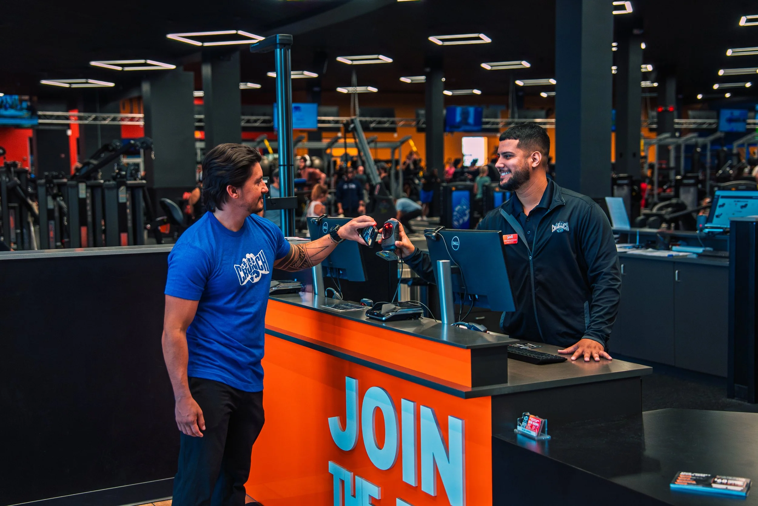 A man and a woman at a retail checkout counter in a gym or fitness center. The woman is smiling while scanning an item, and the man is smiling as he interacts with her. The background shows exercise equipment and people working out.