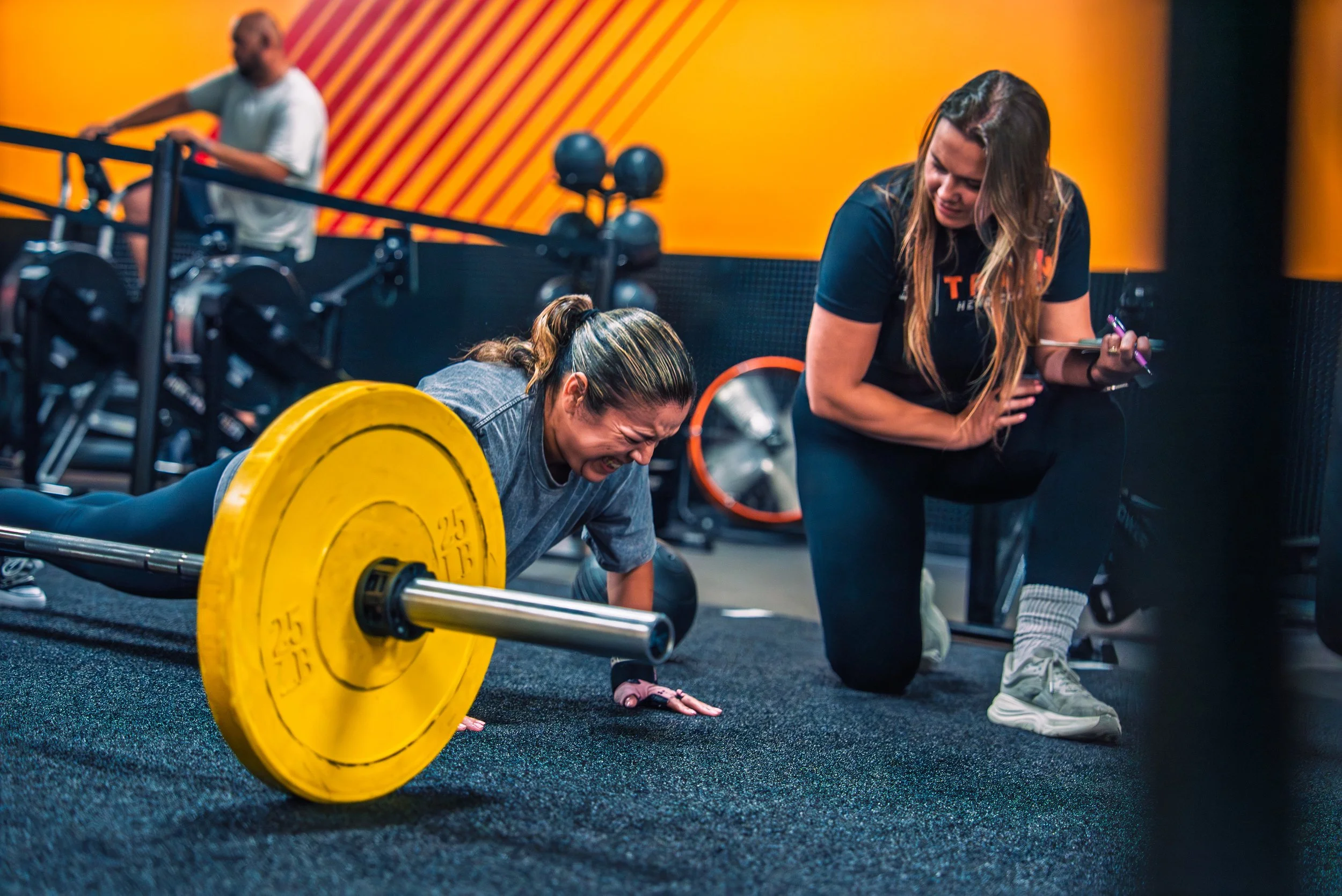 A woman performing a push-up with weights on her back in a gym, with a trainer closely observing and taking notes.