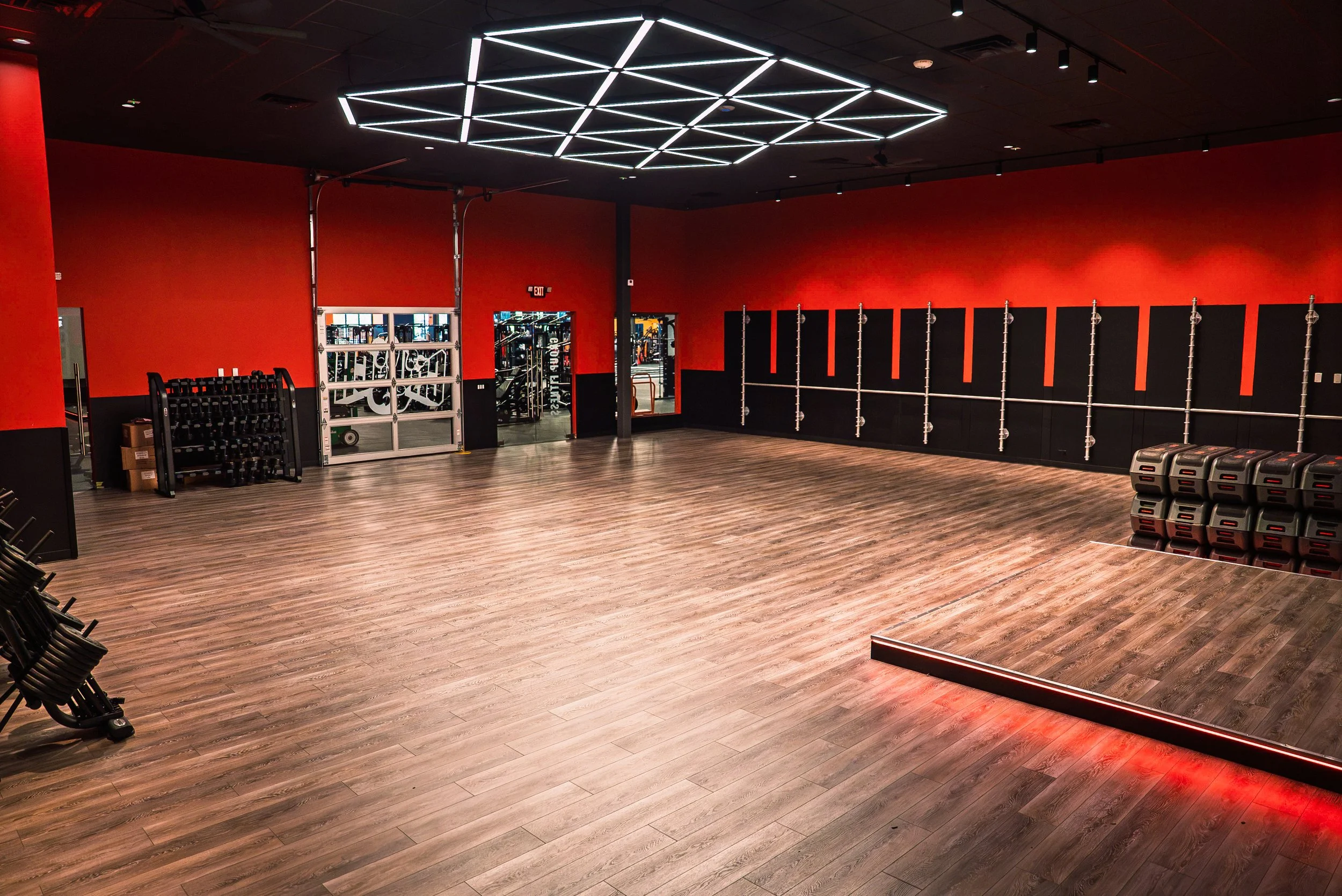 Interior of a gym with black and red walls, wooden floor, and various fitness equipment including weights and medicine balls. The ceiling has a modern geometric light fixture.