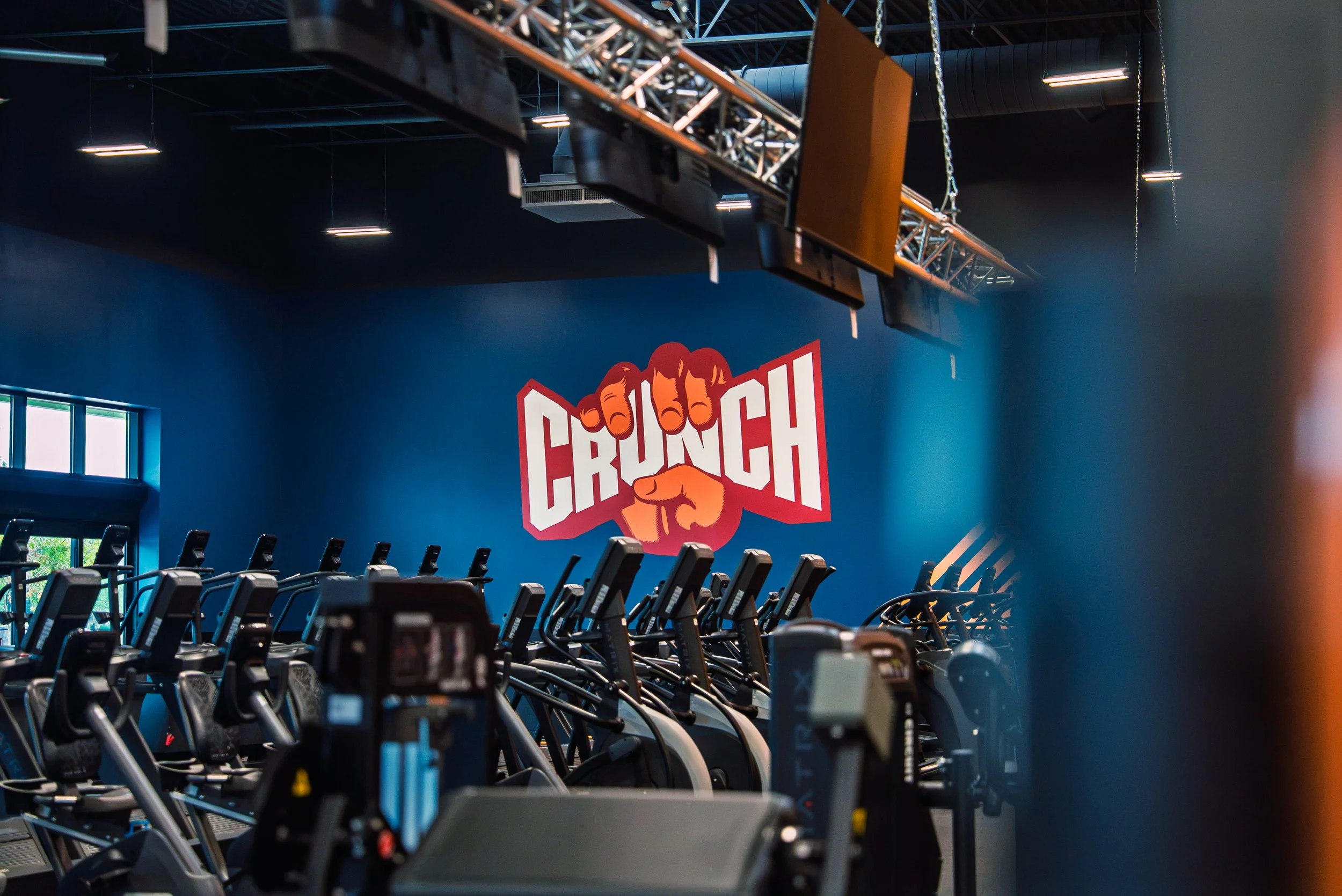 Indoor gym with rows of stationary exercise bikes, a blue wall with a red and white sign that says 'CRUNCH', and ceiling-mounted lighting and equipment.
