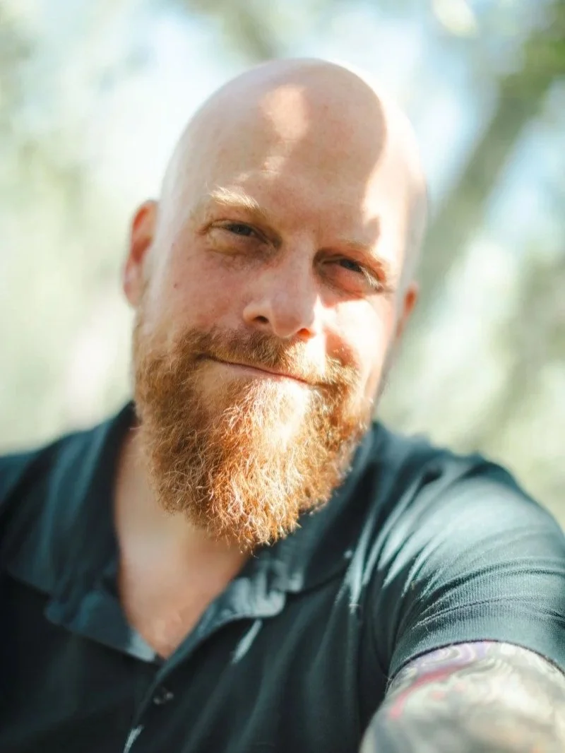 A close-up of a smiling man with a beard, tattoos, and a shaved head outdoors in bright sunlight.