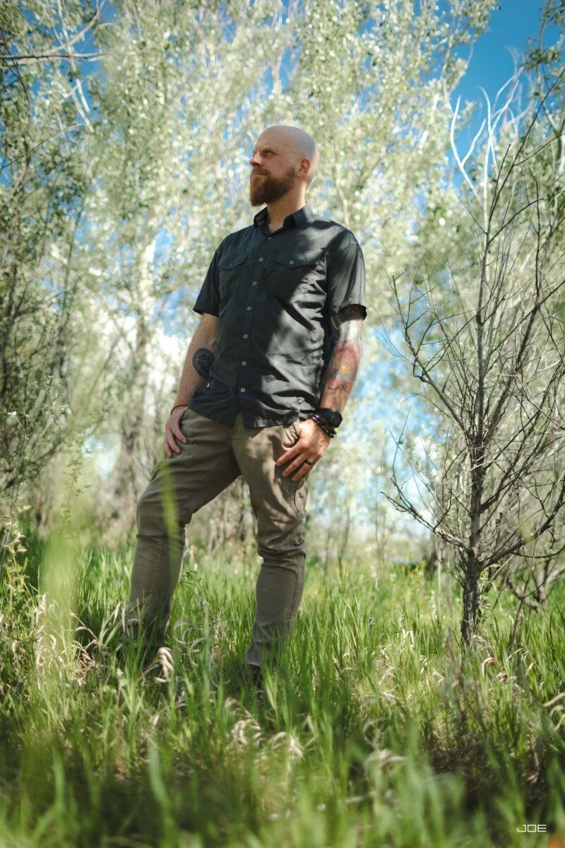 A man with a beard and tattoos standing outdoors among green grass and trees, wearing a black short-sleeve shirt and brown pants, looking off into the distance on a sunny day.