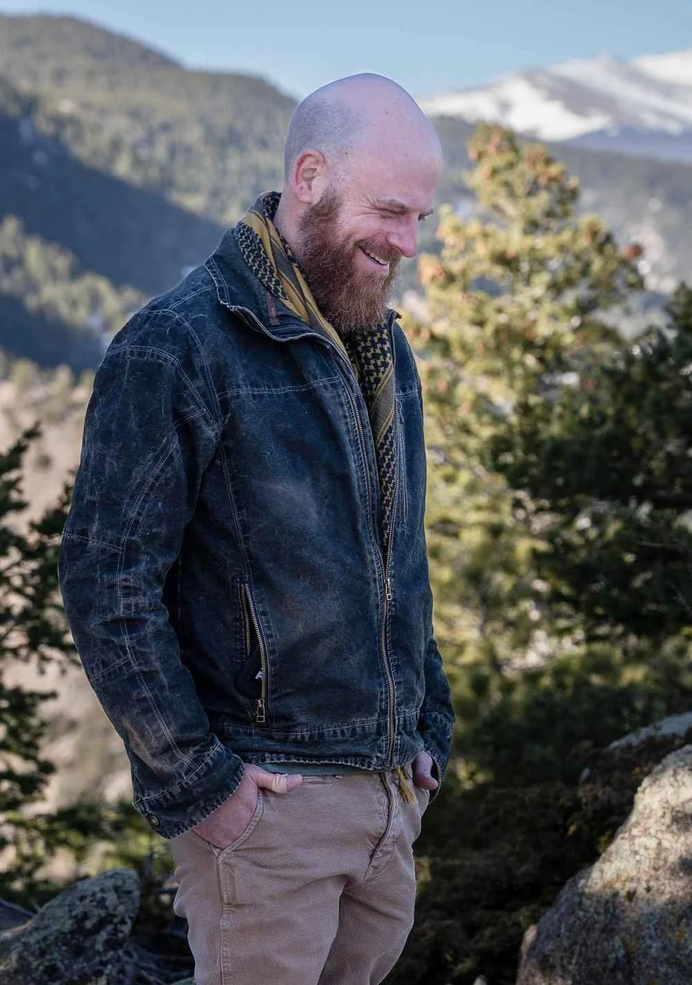 Nicholas Whitaker with beard wearing a denim jacket and scarf standing outdoors near mountains.