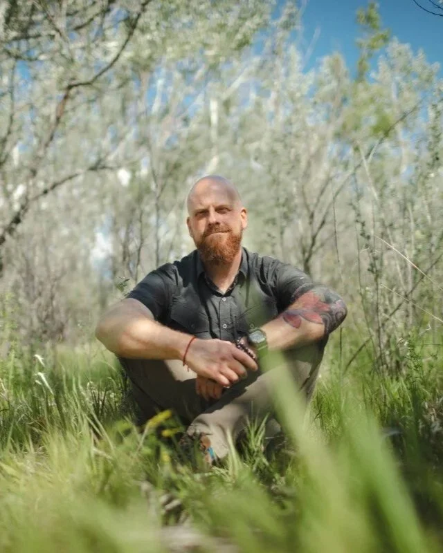 A man sitting cross-legged in a grassy field with trees in the background, holding a knife in his right hand and looking at the camera.