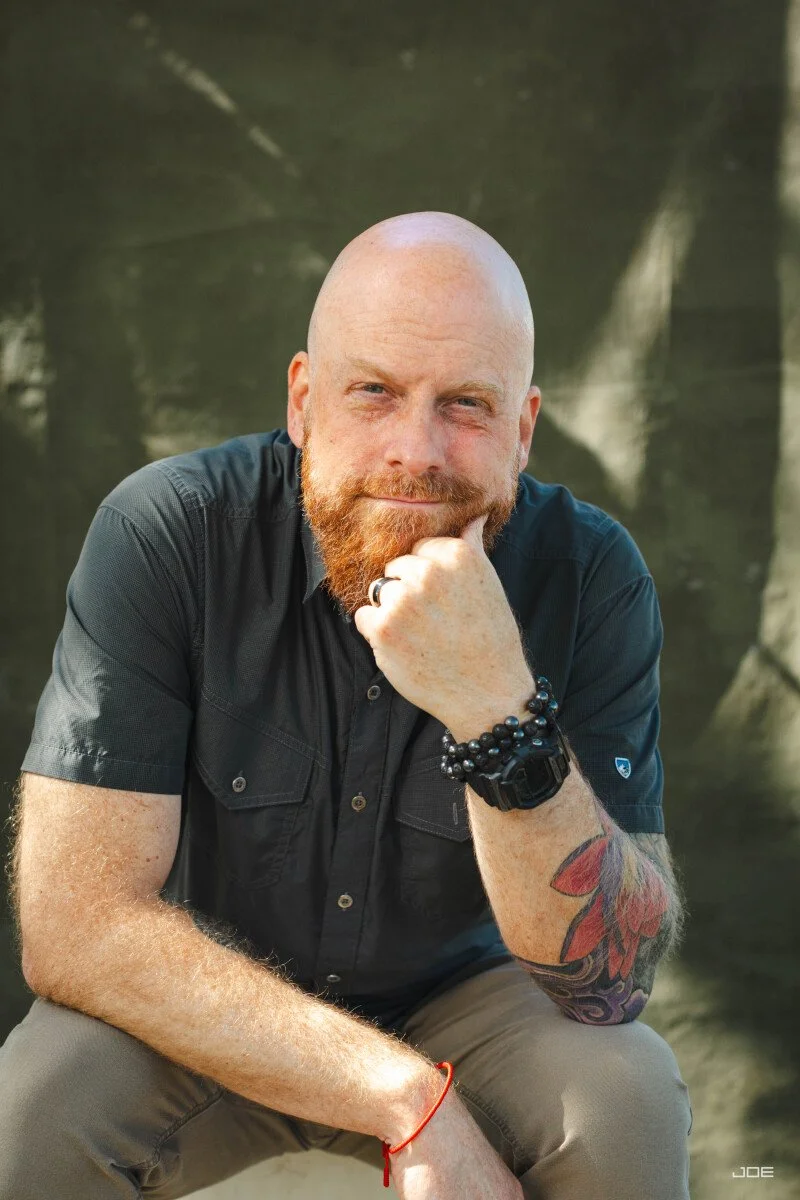 A man with a beard sitting outdoors with a thoughtful expression, wearing a black shirt, beige pants, and accessories including a watch, bracelets, and ring, with a tattoo on his arm and a blurry natural background.