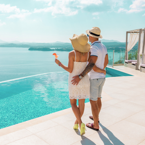 A couple stands by an infinity pool overlooking a lake or sea. The woman is wearing a wide-brimmed hat and yellow heels, the man is wearing a straw hat and shorts. They are embracing and enjoying the view.