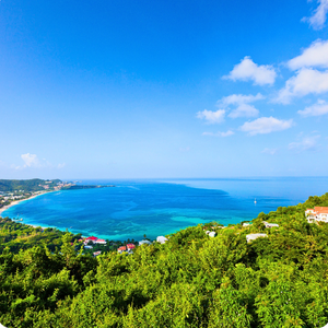 A panoramic view of a coastal bay with clear blue water, surrounded by green hills and a bright blue sky with some clouds.