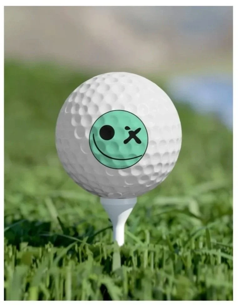 Golf ball with a green smiley face sticker featuring a winking eye, placed on a tee in a grassy area.