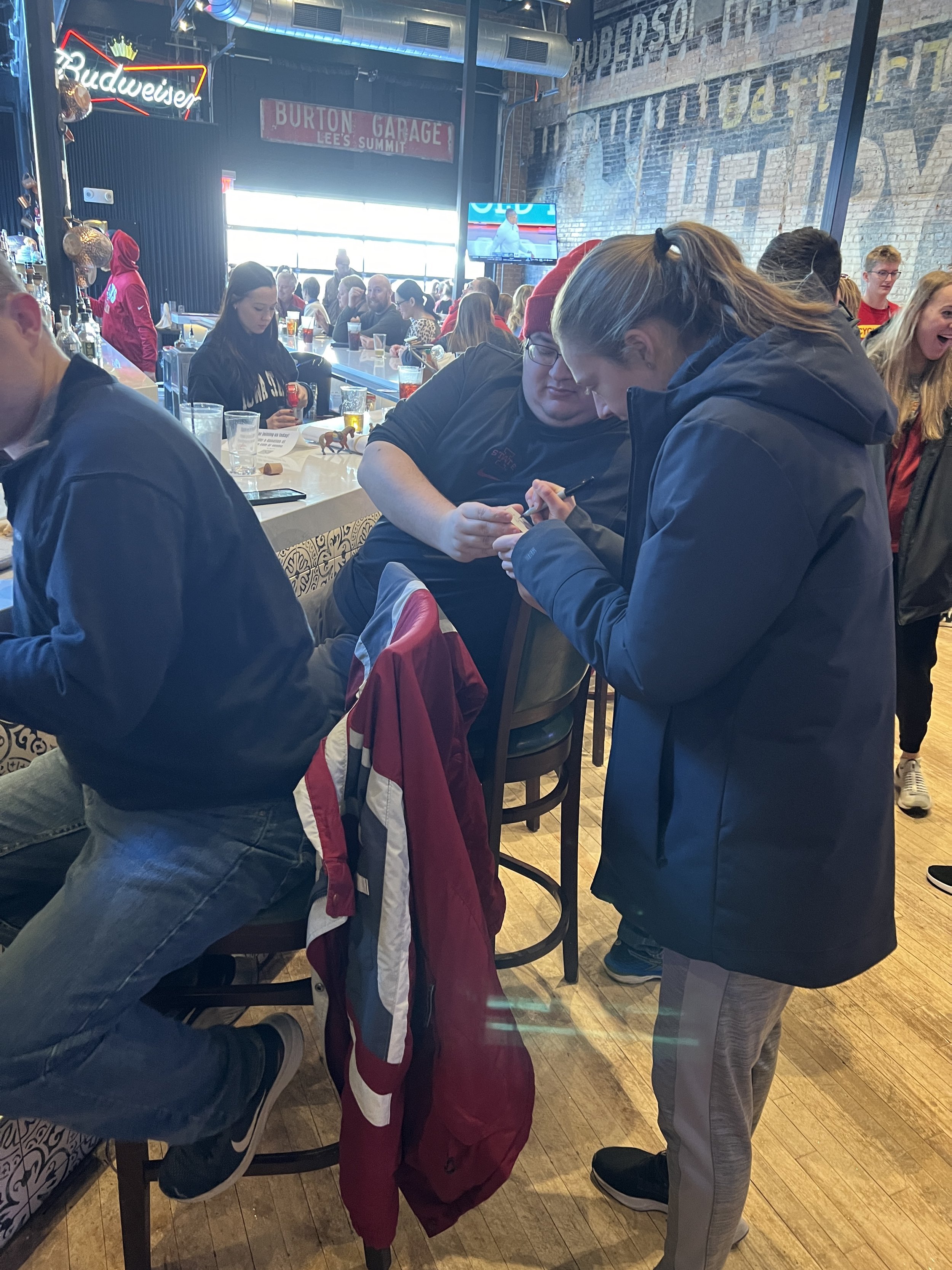 People socializing in a bar with casual seating. "Budweiser" sign and vintage-style decor are visible. Some individuals are seated at the counter, while others are standing, engaged in conversation.