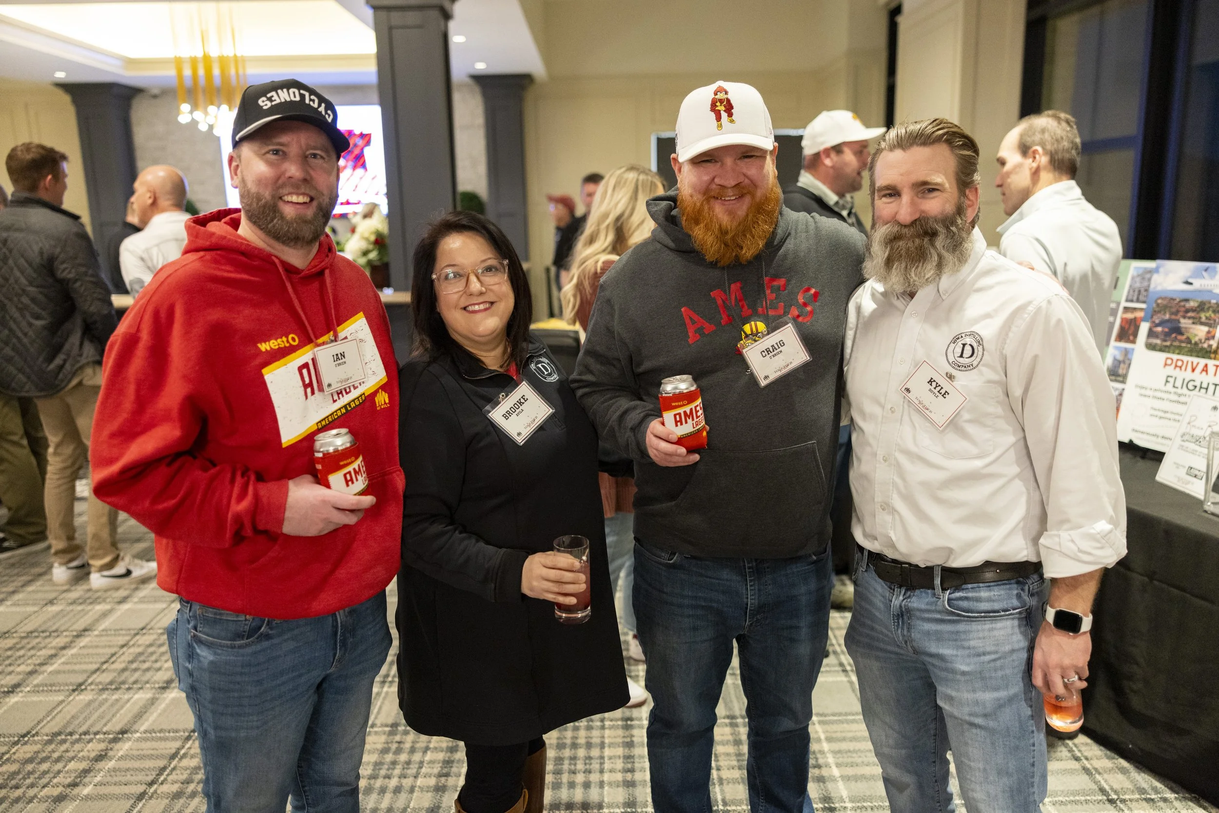 Group of four people posing at an event, holding drinks with name tags. One person is wearing a "Cyclones" hat, another wearing a red hoodie with "Ames" branding. Background shows other attendees and promotional materials.