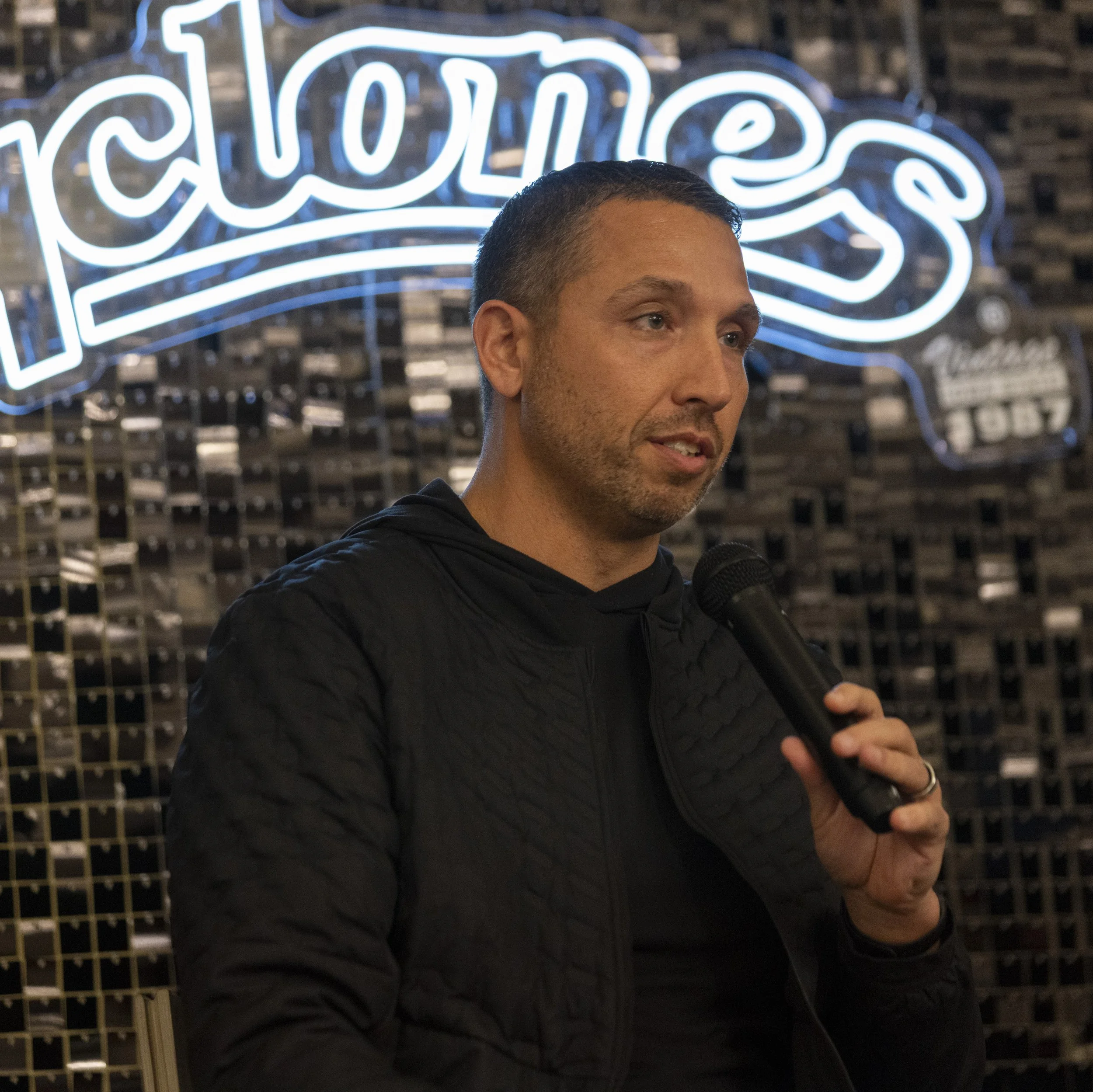 A man holding a microphone in front of a sign with "cyclones" in cursive neon lettering.