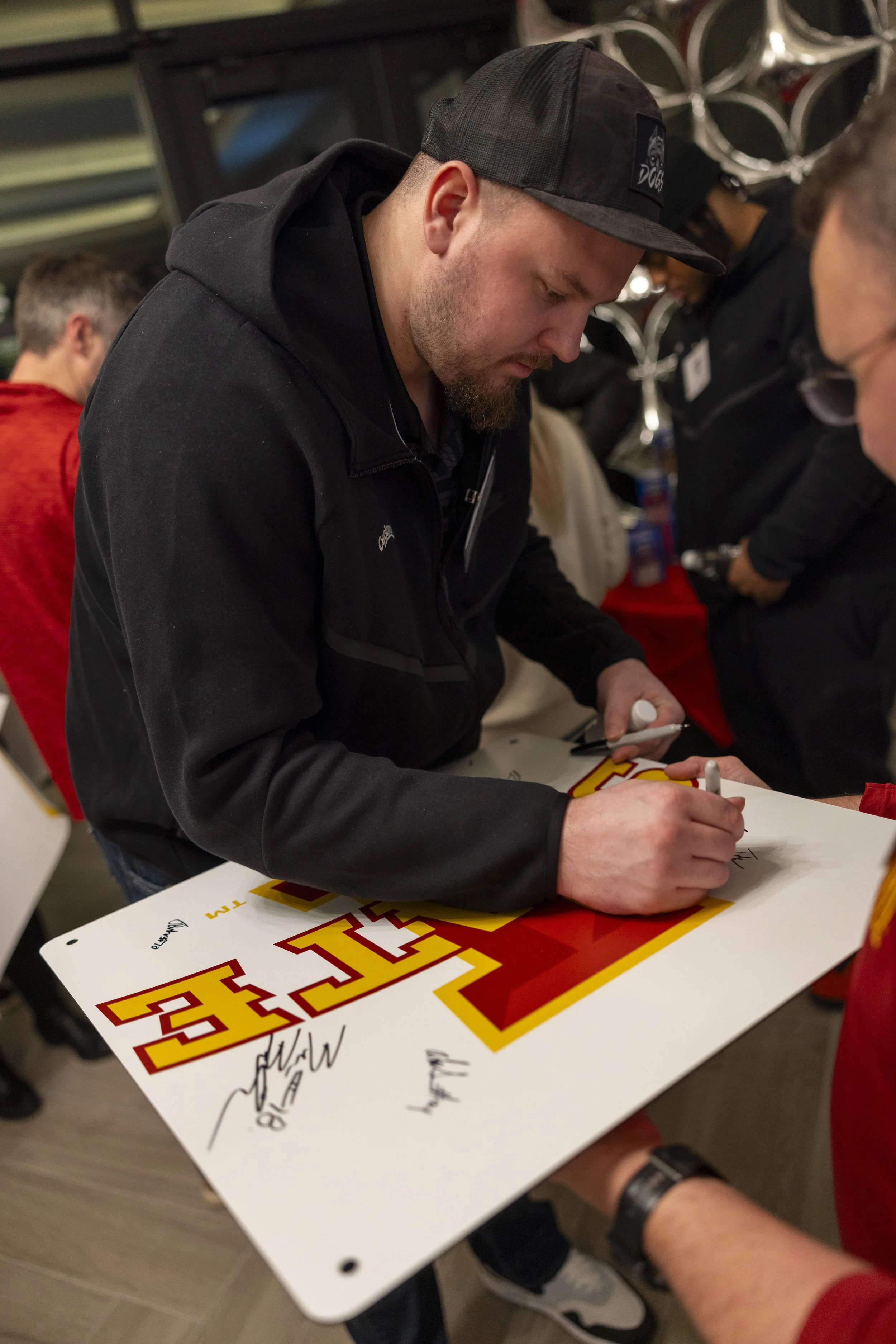 A man signing a large board with a sports team logo, wearing a black jacket and cap, surrounded by people indoors.