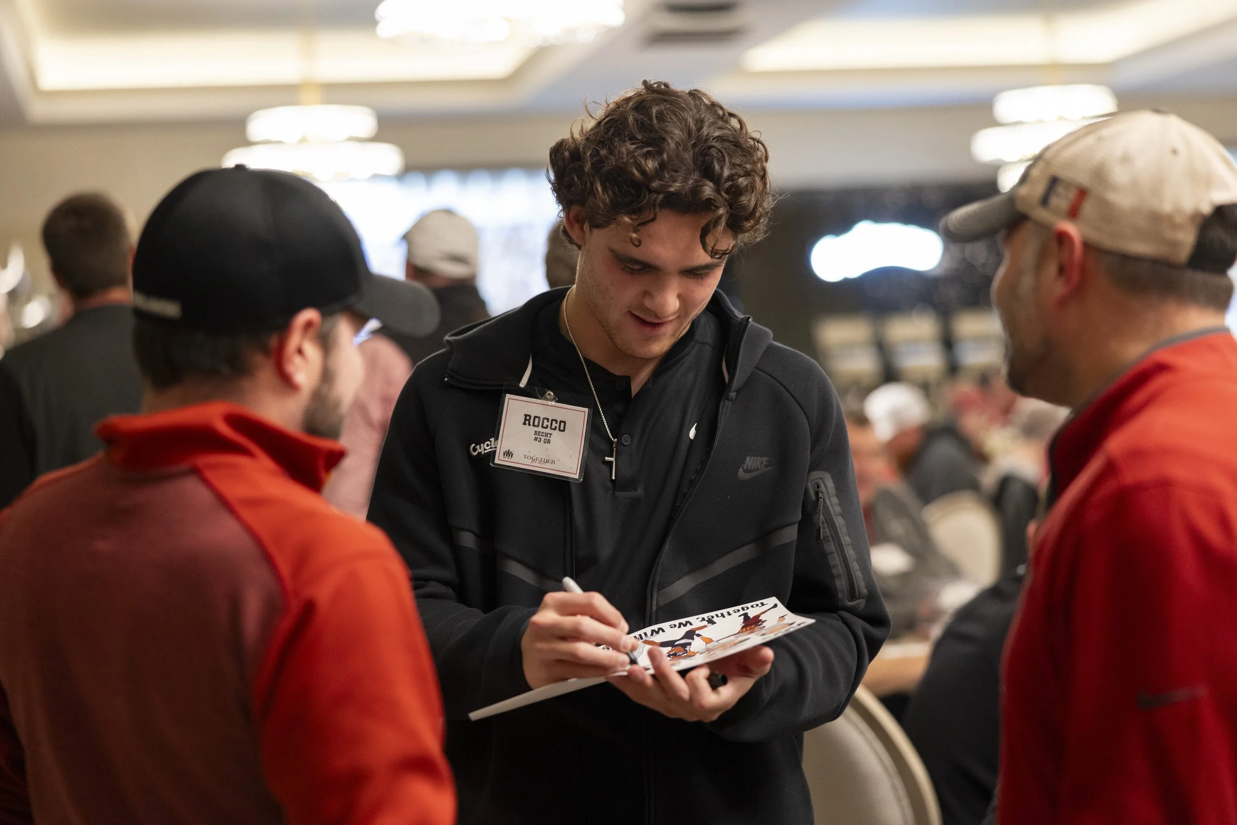 A man writing on a notepad in a crowded room with two other people engaged in conversation.