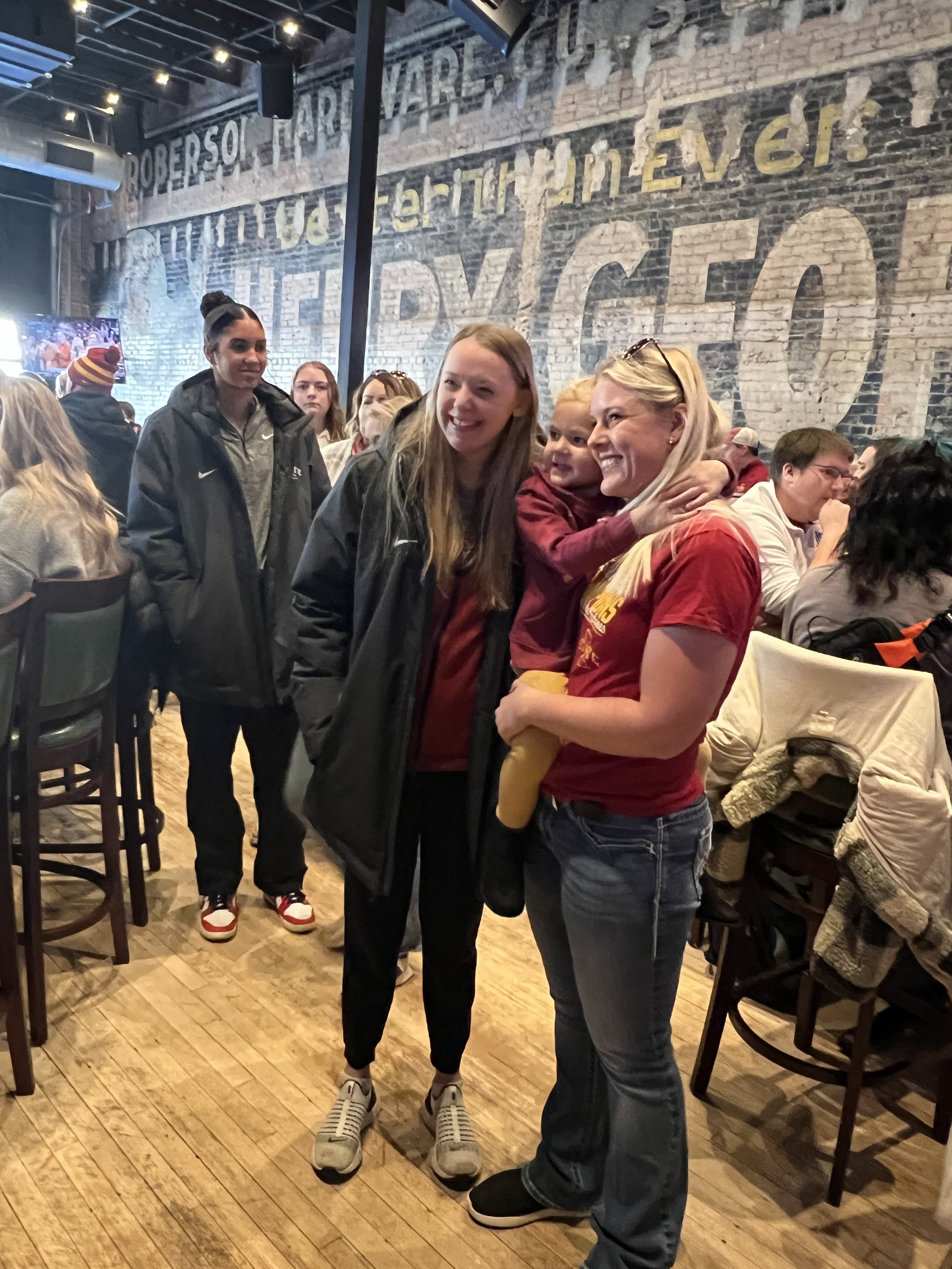 Group of people in a casual restaurant with brick walls, including a smiling woman holding a baby, other adults standing nearby, and diners seated in the background.