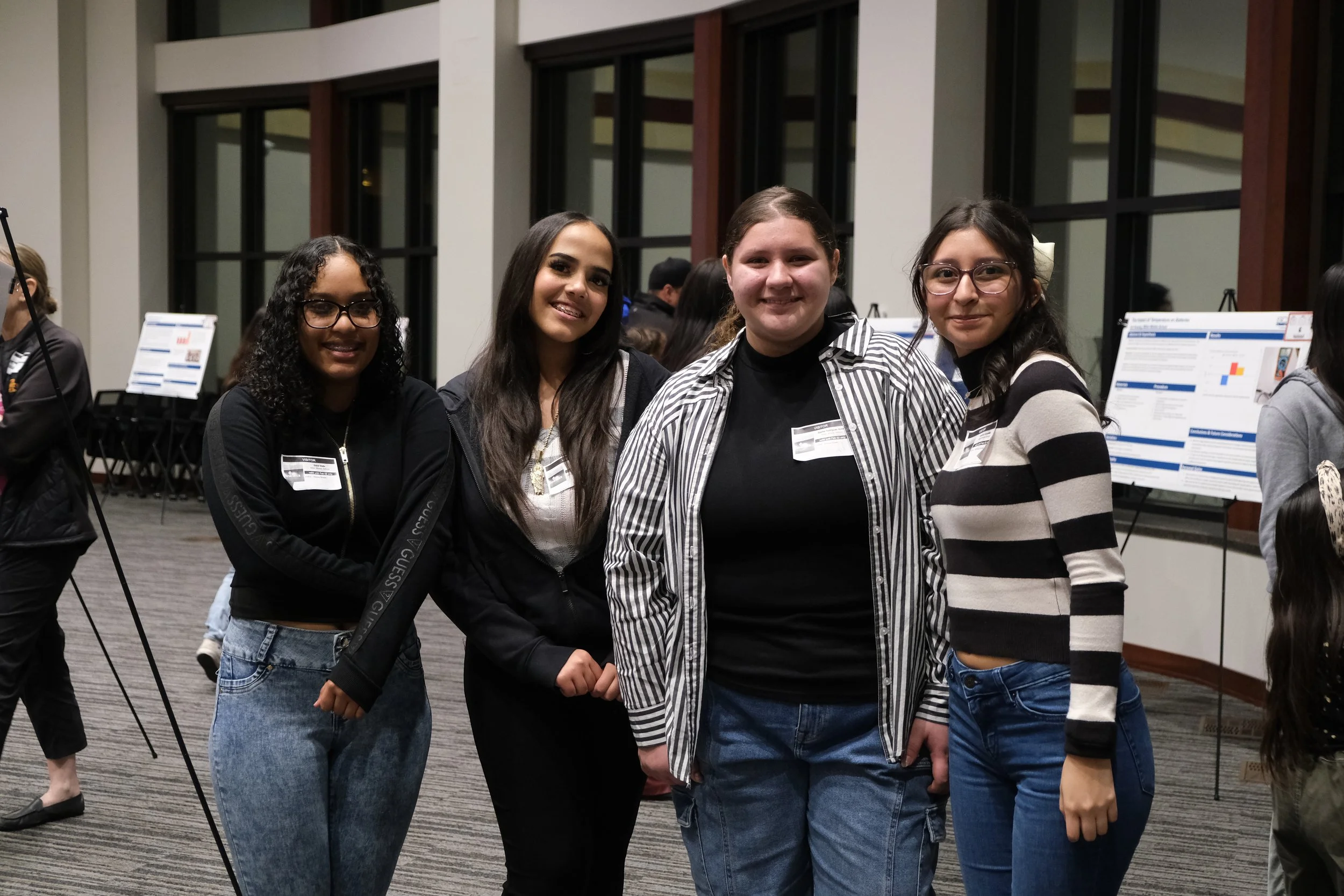 Group of four women smiling at an indoor event with presentation boards in the background.