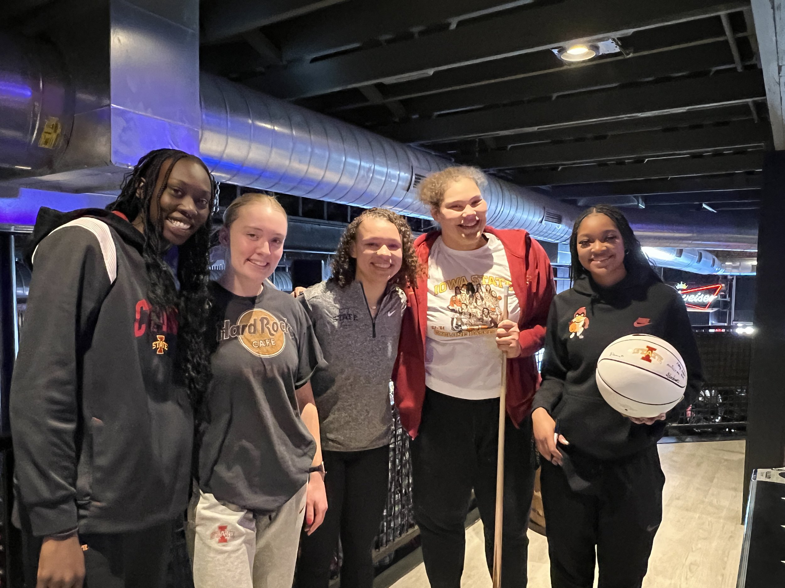 Group of five women smiling indoors, with one holding a basketball.