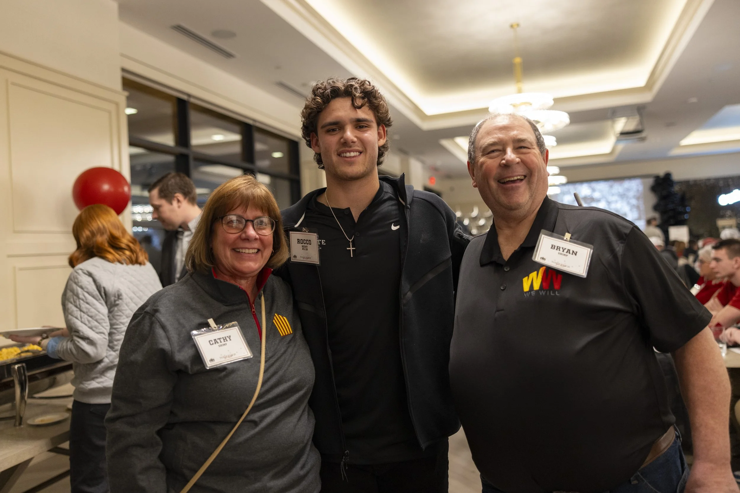 Three people smiling and posing at an indoor event with name tags, surrounded by a lively atmosphere.
