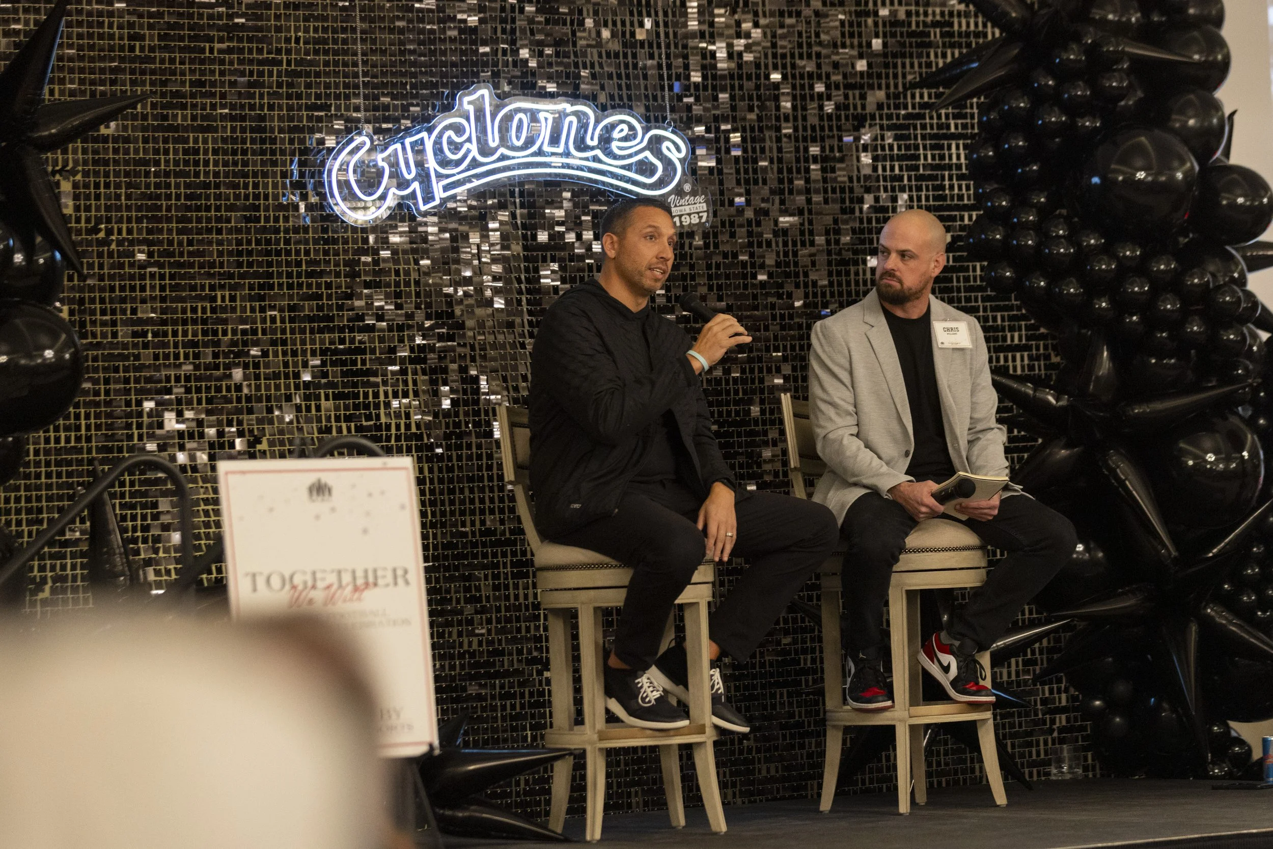 Two people sitting on high chairs on a stage surrounded by black balloons; one holds a microphone, with the word 'Cyclones' illuminated behind them.