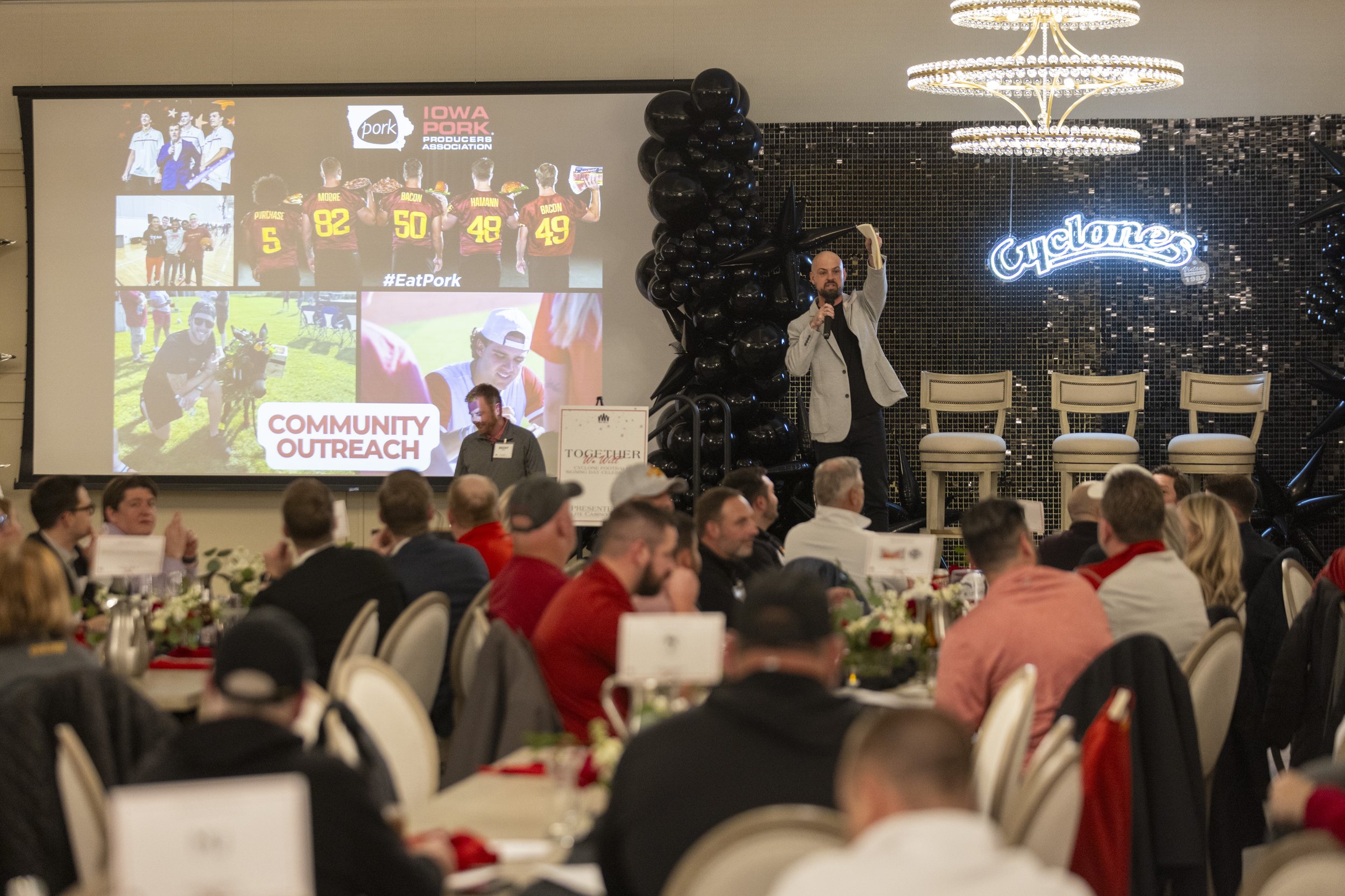 A speaker on stage at a community outreach event with Iowa Pork Producers Association branding, addressing an audience seated at tables. The background features images of sports players and a neon sign reading 'Cyclones.'