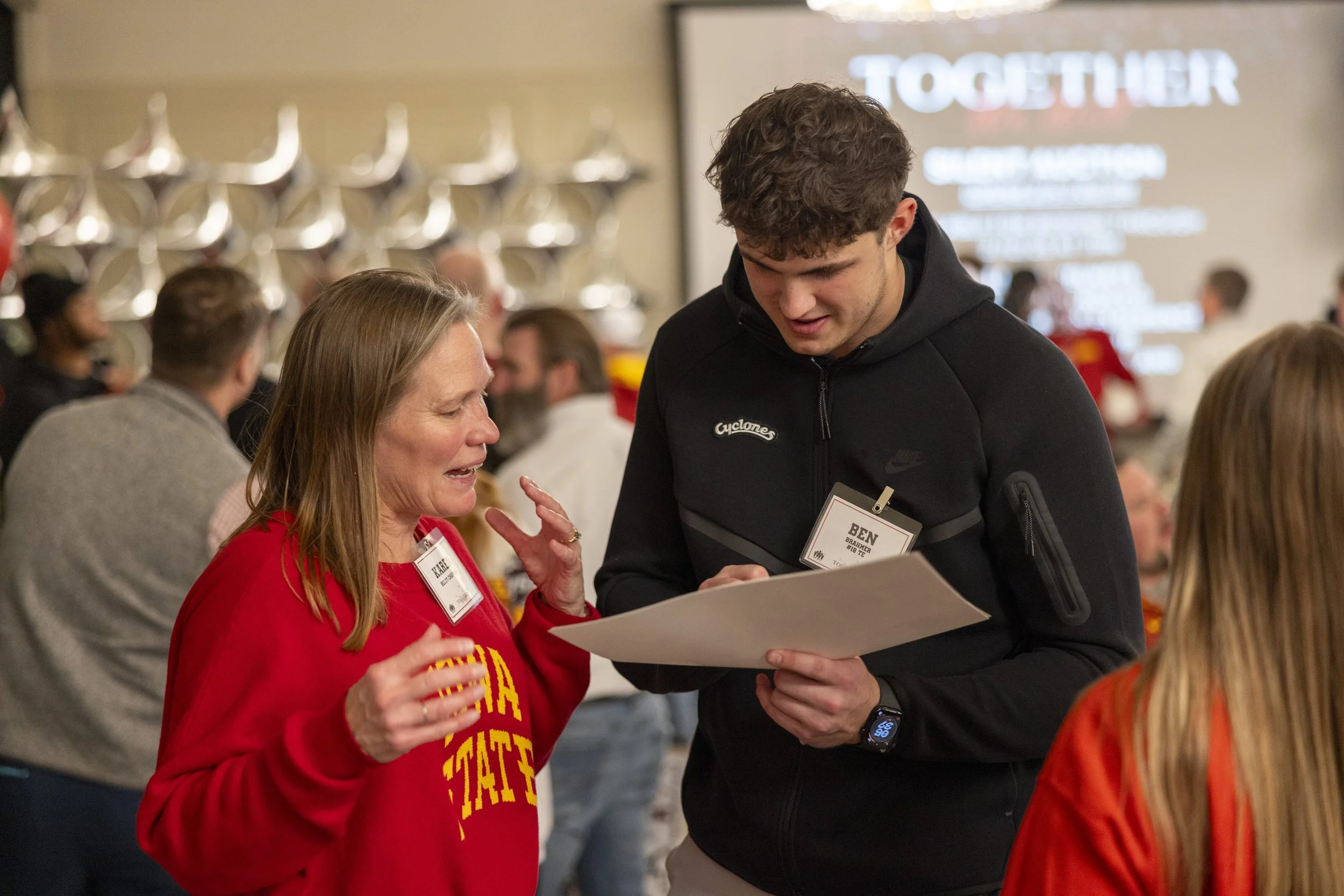 People in a room at an event, with name tags; a woman gesturing and talking to a man holding papers, background presentation screen.