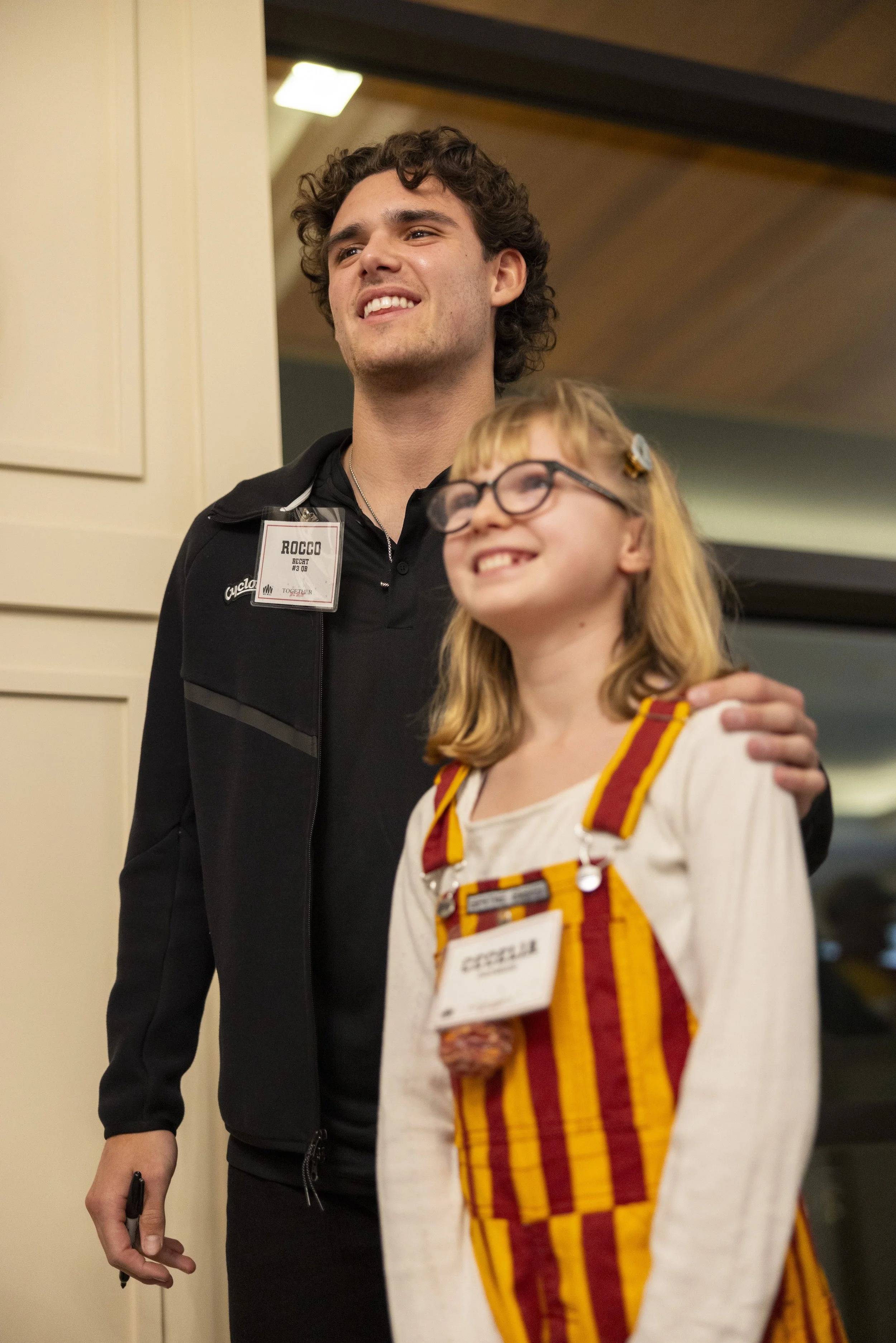 A young man and girl smiling indoors, wearing name tags in a conference setting.