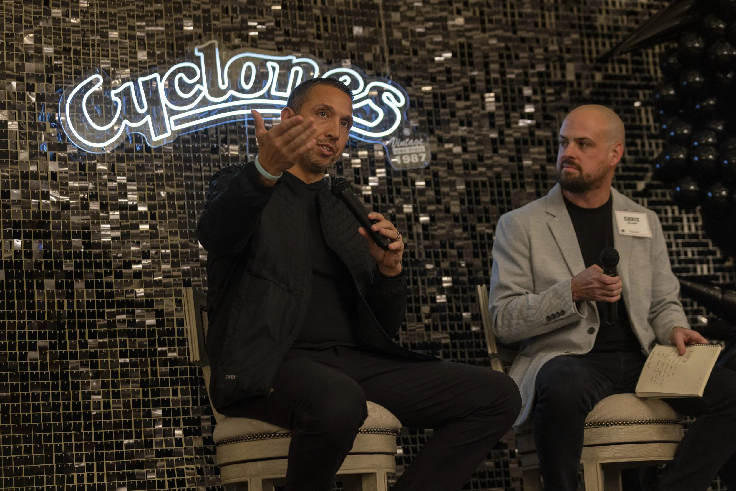 Two men sitting on chairs with microphones, one gesturing, in front of a wall with a "Cyclones" sign.