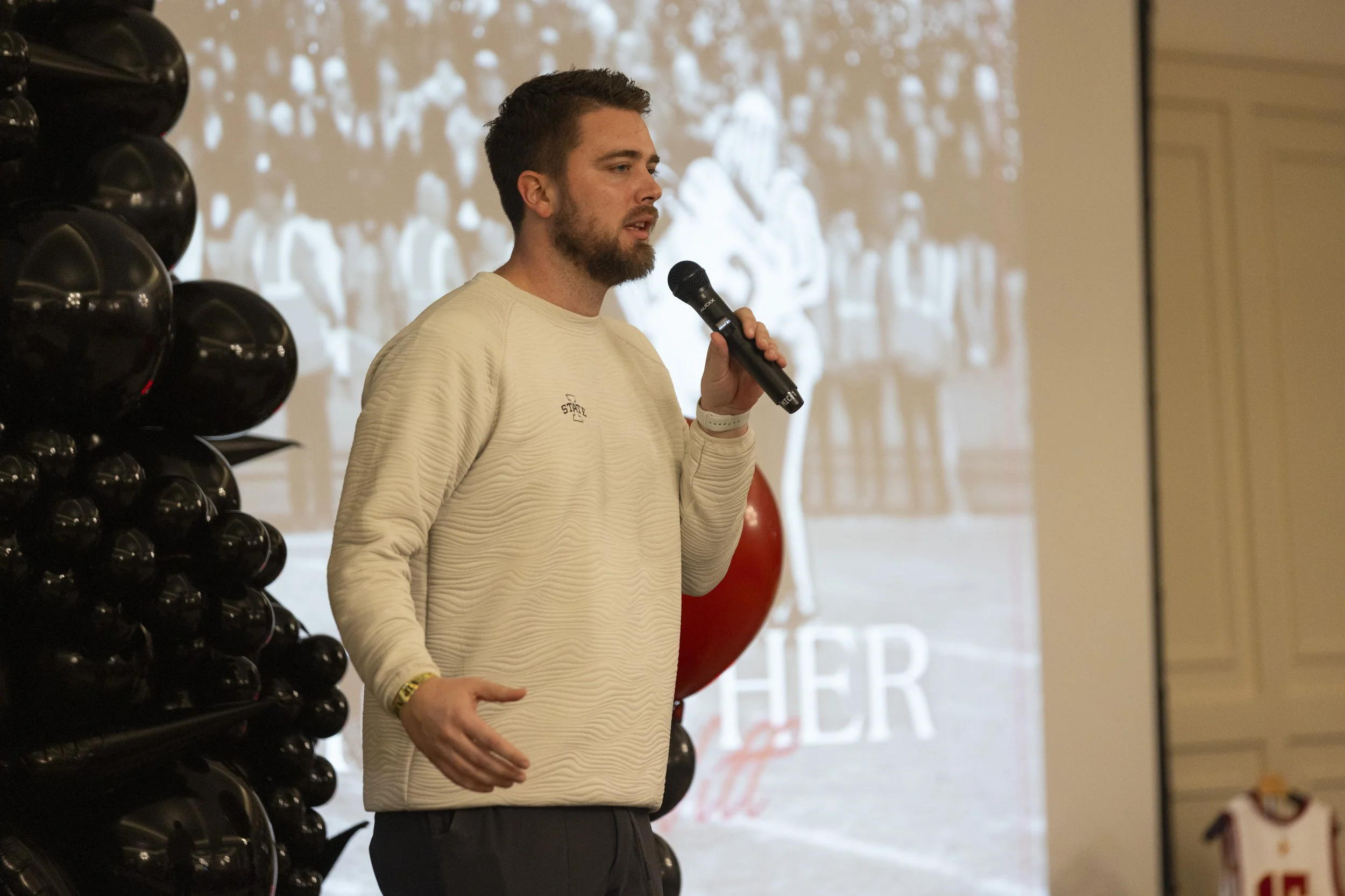Man speaking into a microphone at an indoor event, standing near black balloons with a blurred background featuring an image of a crowd.