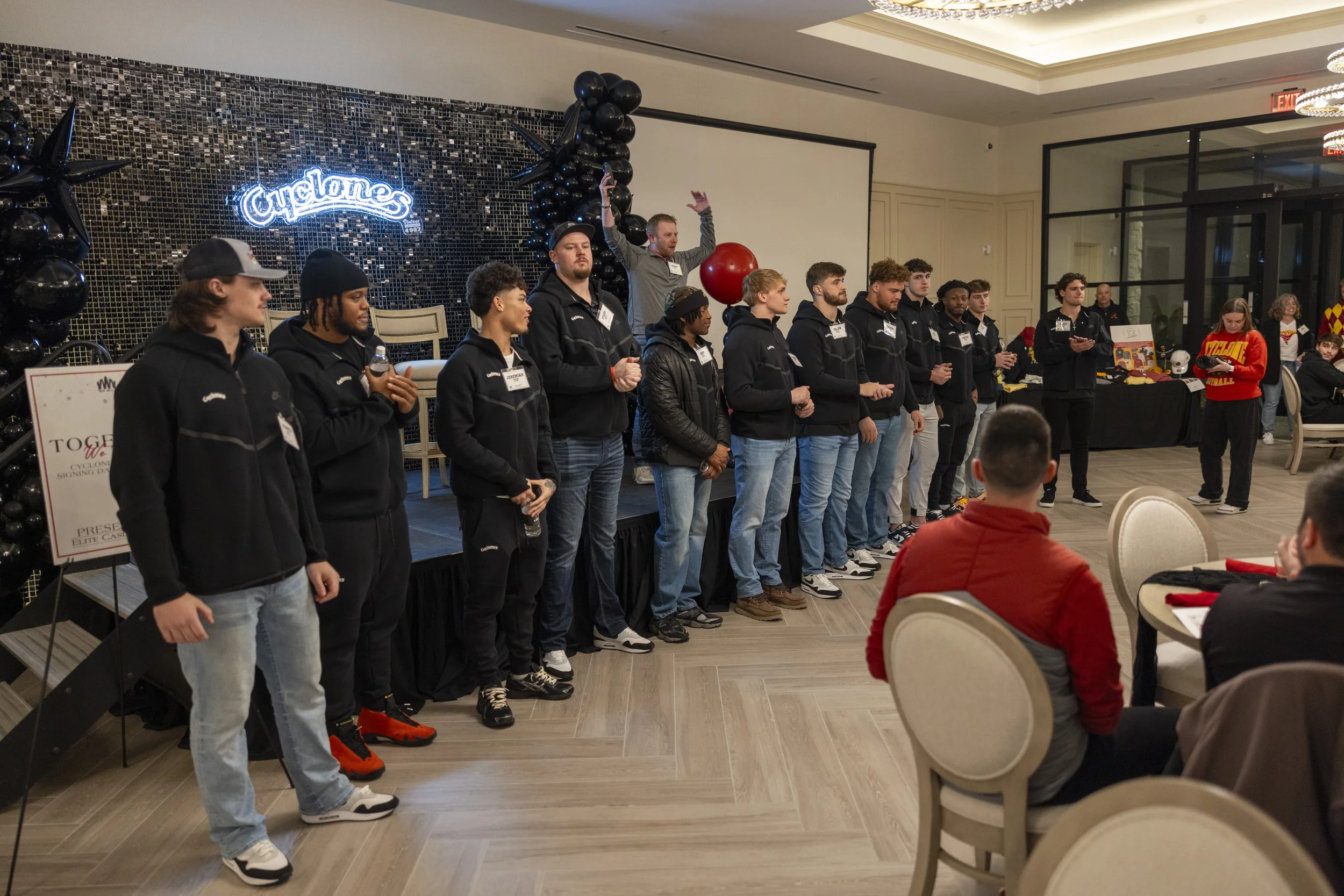 A group of people standing on a stage in a room with a 'Cyclones' sign. They are wearing casual attire with name tags, gathered for an event. Seated attendees are watching. Decor includes black balloons and a mosaic backdrop.