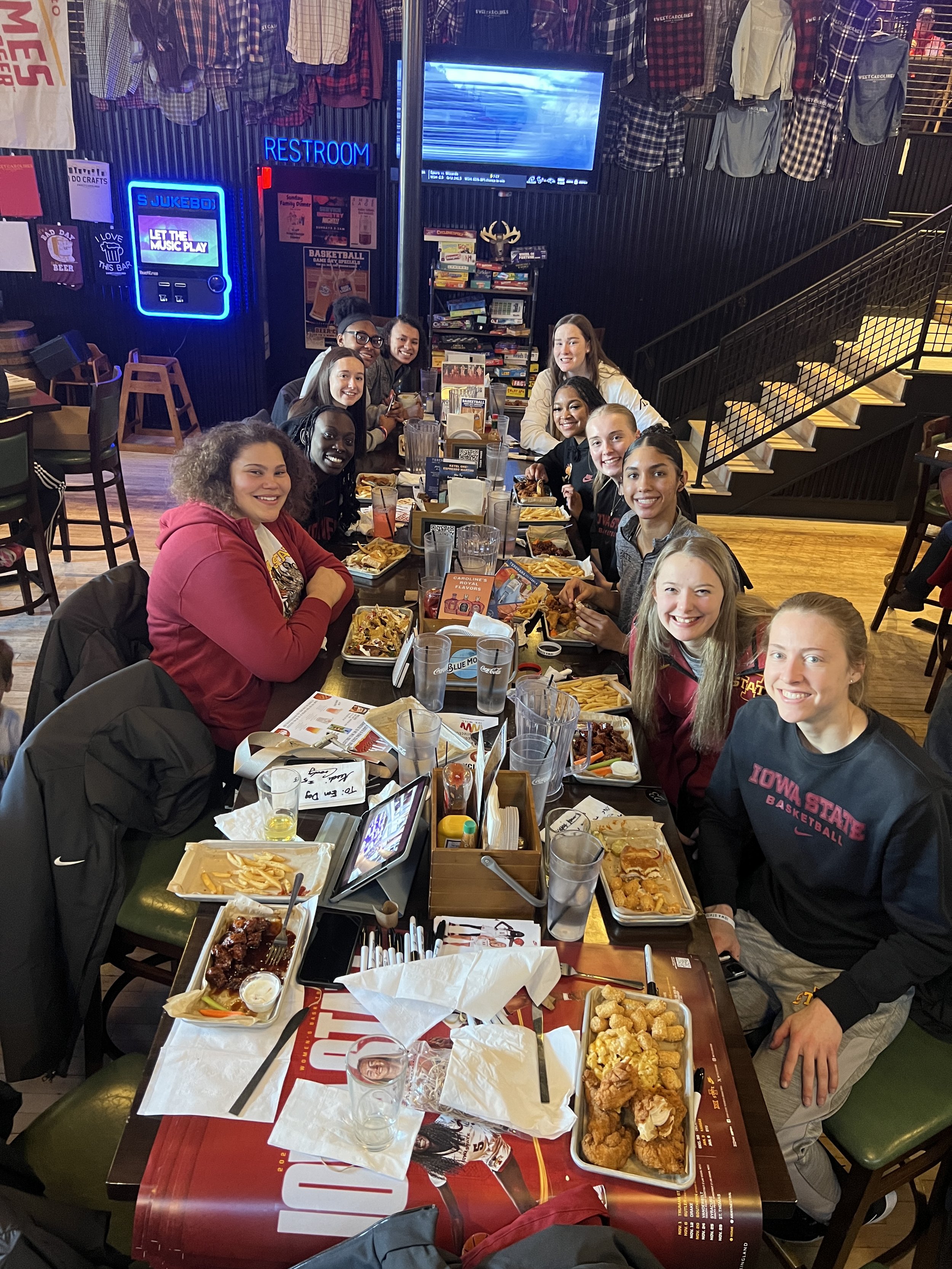 Group of people sitting at a restaurant table with food, including fries and chicken, at a sports-themed venue. Walls decorated with sports jerseys and memorabilia.
