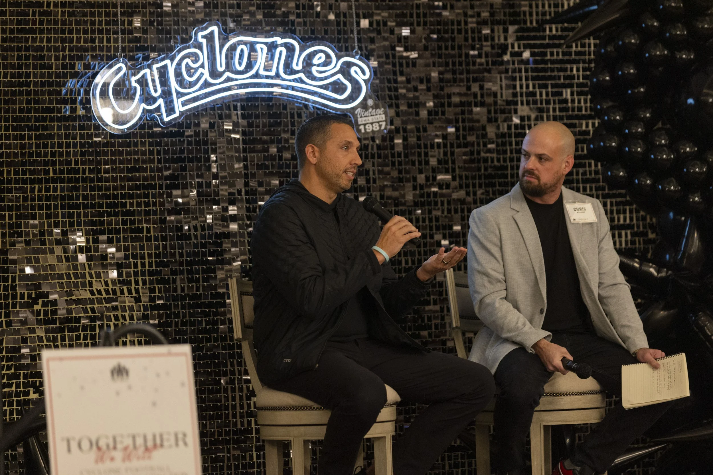 Two men seated on chairs, one speaking into a microphone, with a 'Cyclones' sign in the background and a sign reading 'Together' partially visible in the foreground.