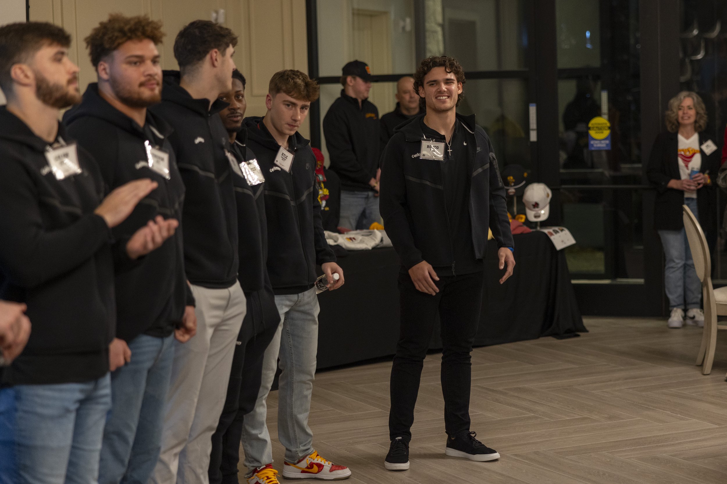 Group of people standing indoors, wearing black jackets with name tags, in a social setting. A table with hats and other items is visible in the background.