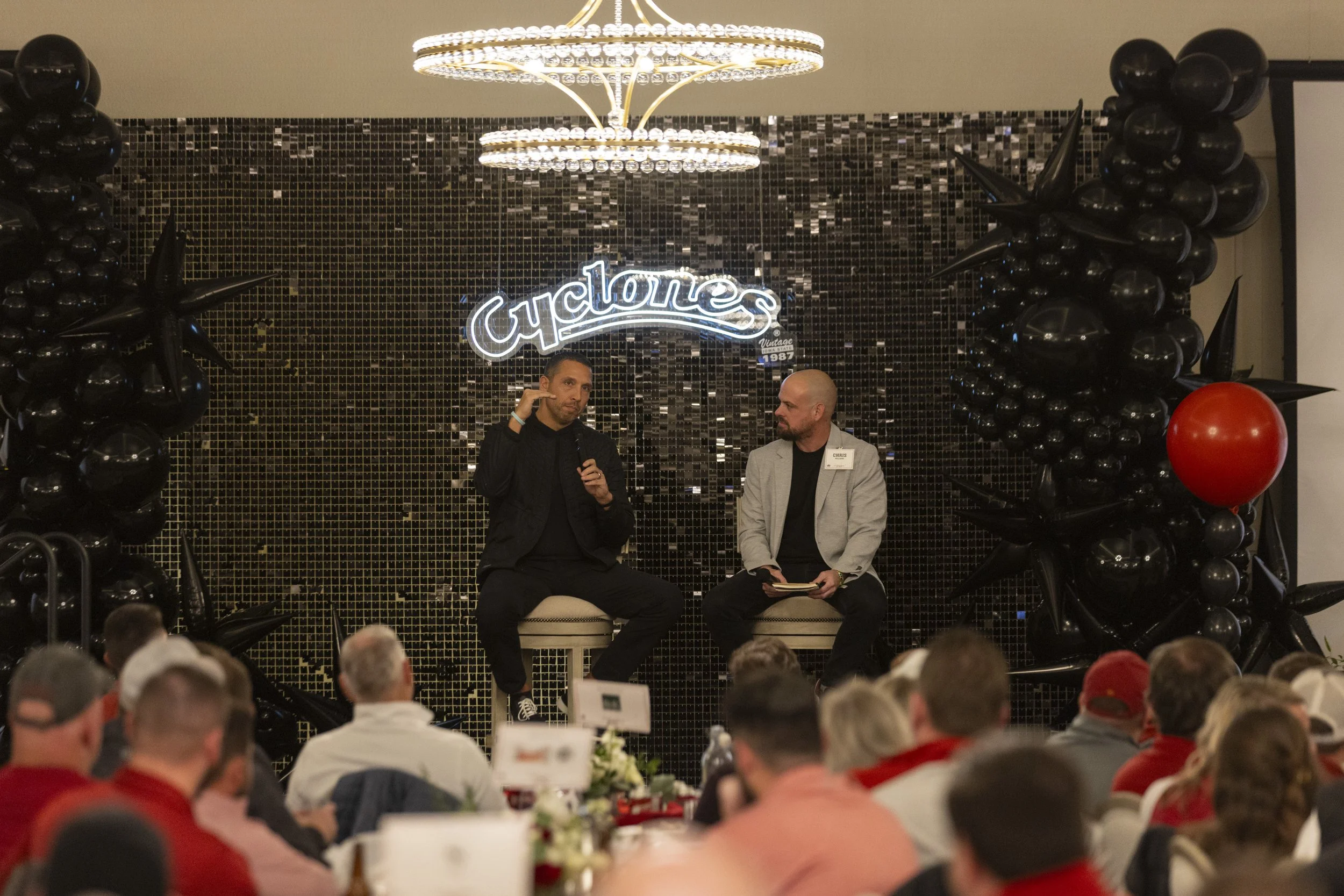 Two people seated on stage in front of an audience, with a "Cyclones" sign and black balloon decorations.